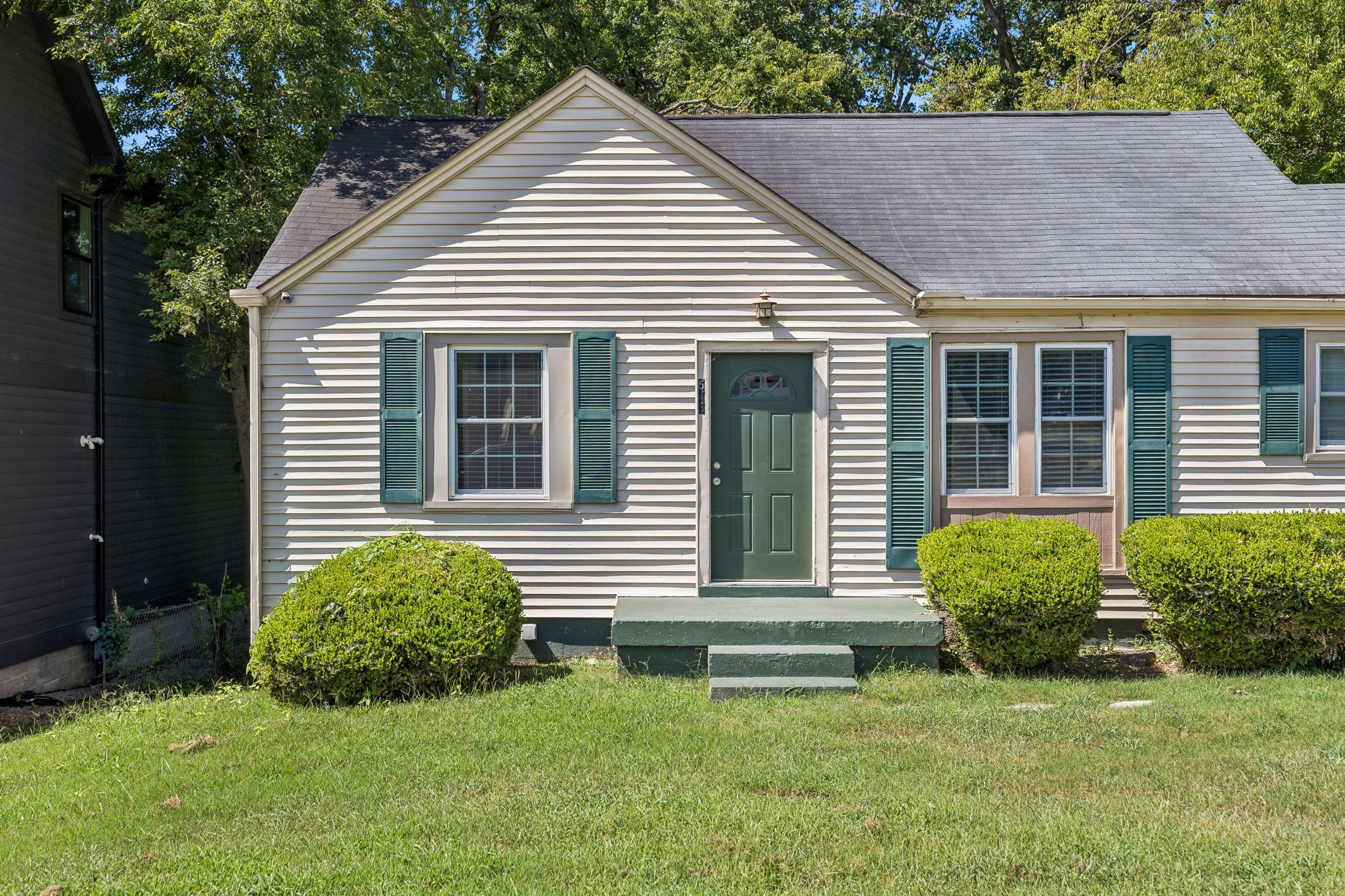 Single-story house with teal shutters and green front door, trimmed shrubs, and a grassy front lawn.