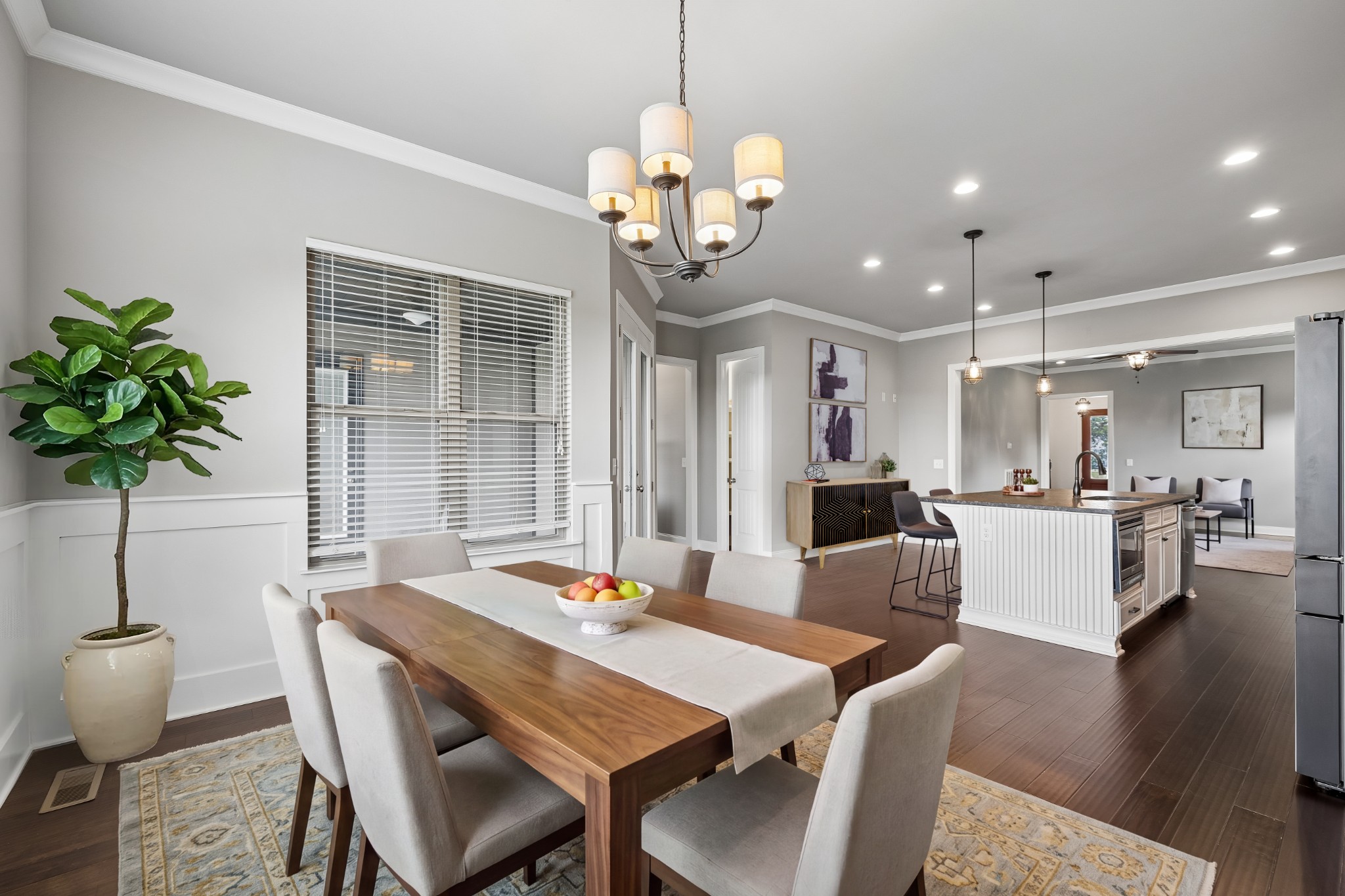 Dining room with a long wooden table and chandelier opening into a kitchen with a white island.