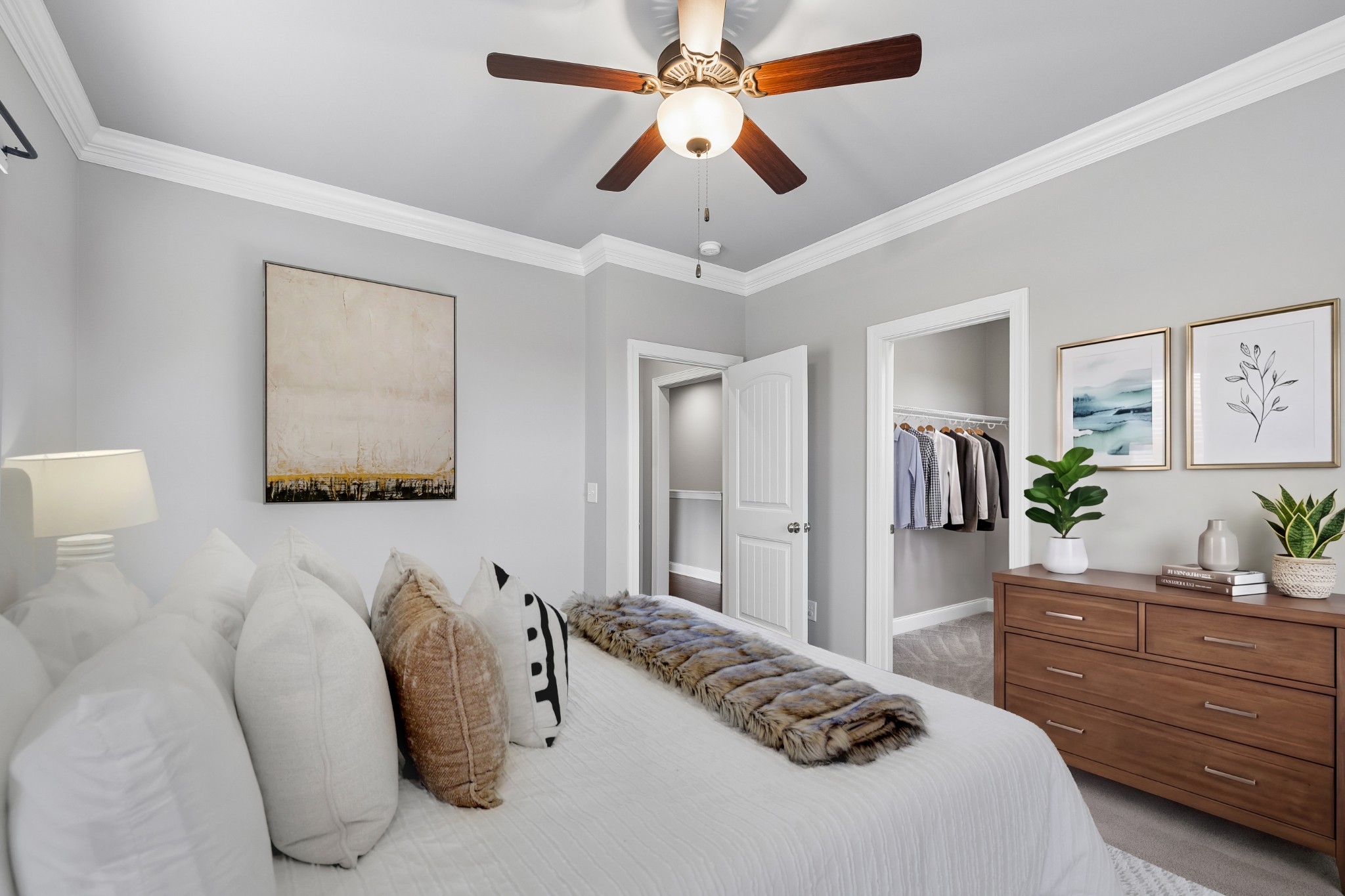 Staged bedroom with a white bed, wooden dresser, ceiling fan, and open closet.