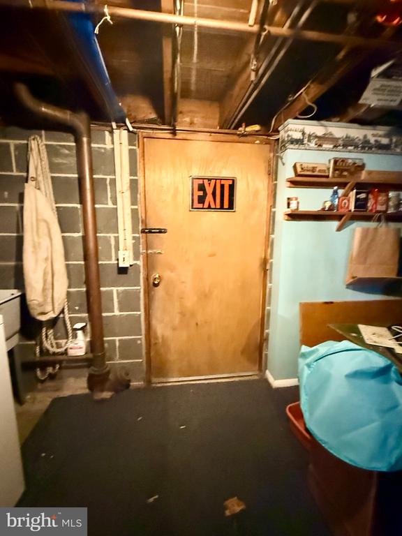 Unfinished basement with exposed ceiling joists, cinder-block walls, pipes, and a wooden exit door.