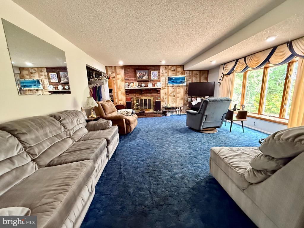 Furnished living room with blue carpet, brick fireplace, and large windows overlooking trees.