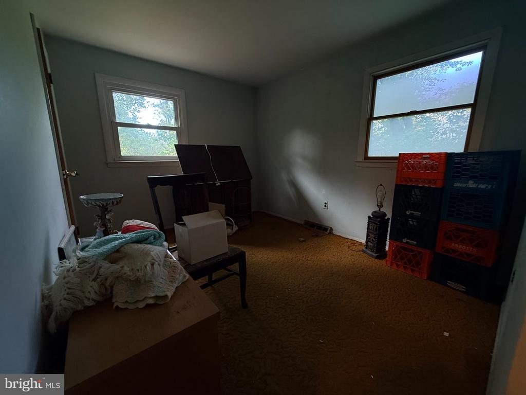 Interior room with brown carpet, two windows, stacked crates, and scattered belongings.