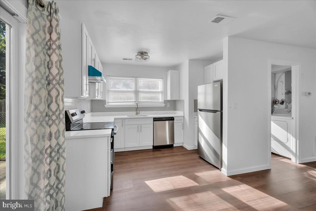 Bright kitchen with white cabinets, stainless steel appliances, and wood-look flooring; laundry area visible through doorway.