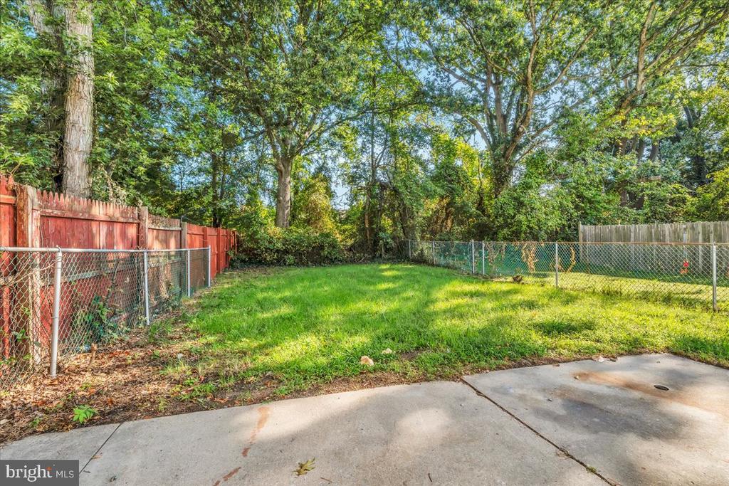 Fenced backyard with a grassy lawn, mature trees, and a concrete patio.