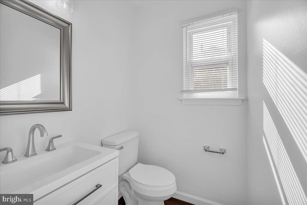 Bright bathroom with a white vanity, framed mirror, toilet, and sunlight through window blinds.