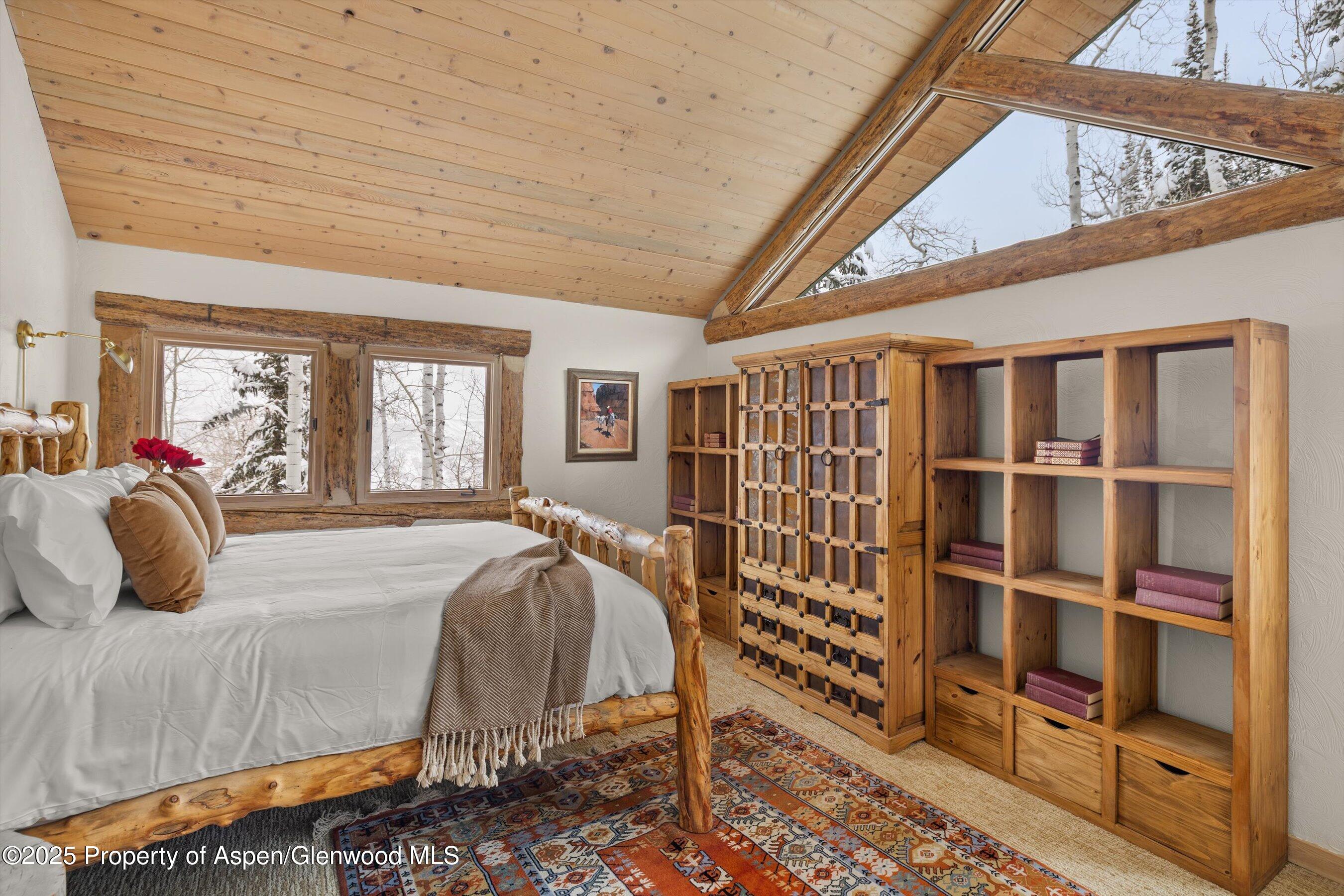 Rustic bedroom with a log bed, vaulted wood ceiling, and windows overlooking snowy trees.