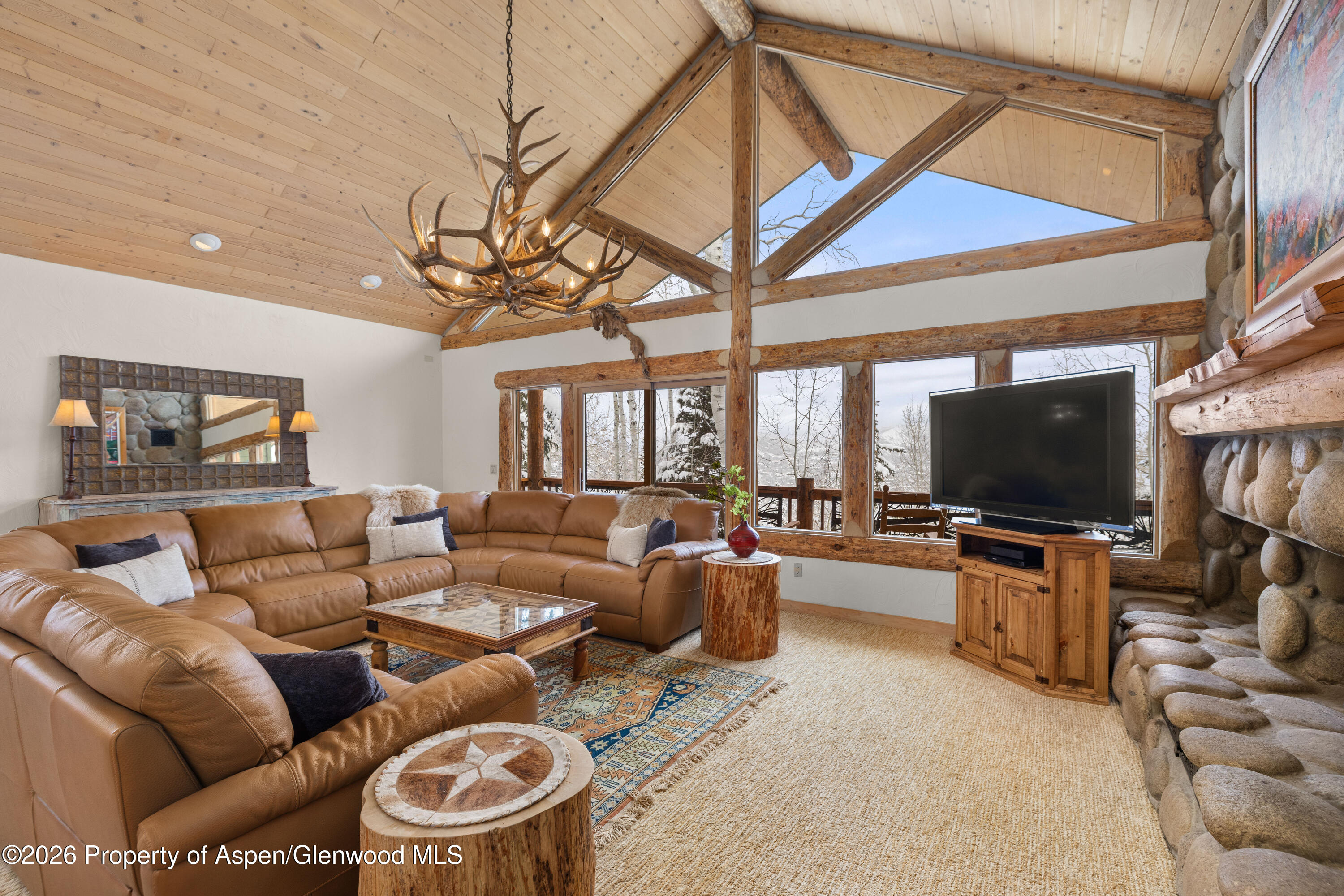 Living room with a leather sectional, vaulted timber ceiling, antler chandelier, and stone fireplace.