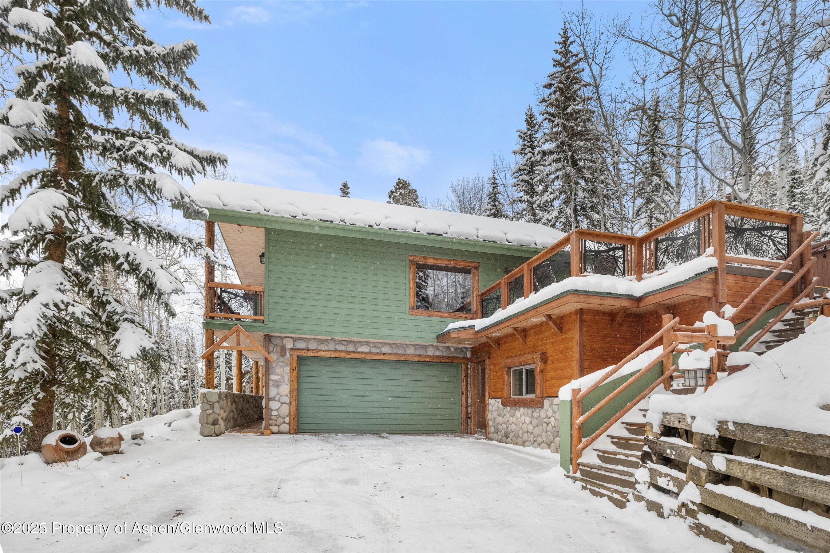 Green mountain home with a two-car garage and raised wooden deck, surrounded by snow-covered trees.