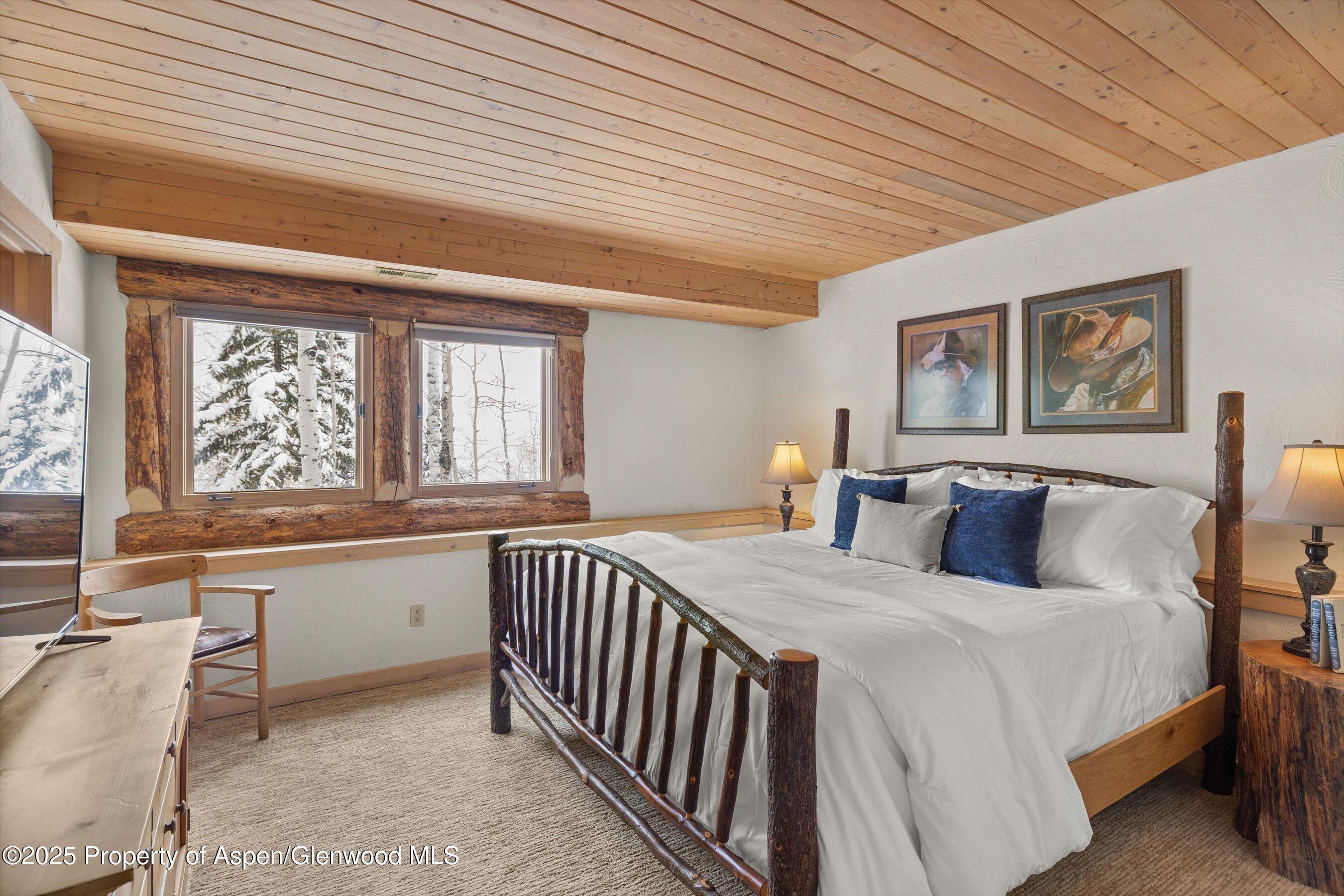 Rustic bedroom with a log-frame bed, vaulted wood ceiling, and snowy trees outside the windows.