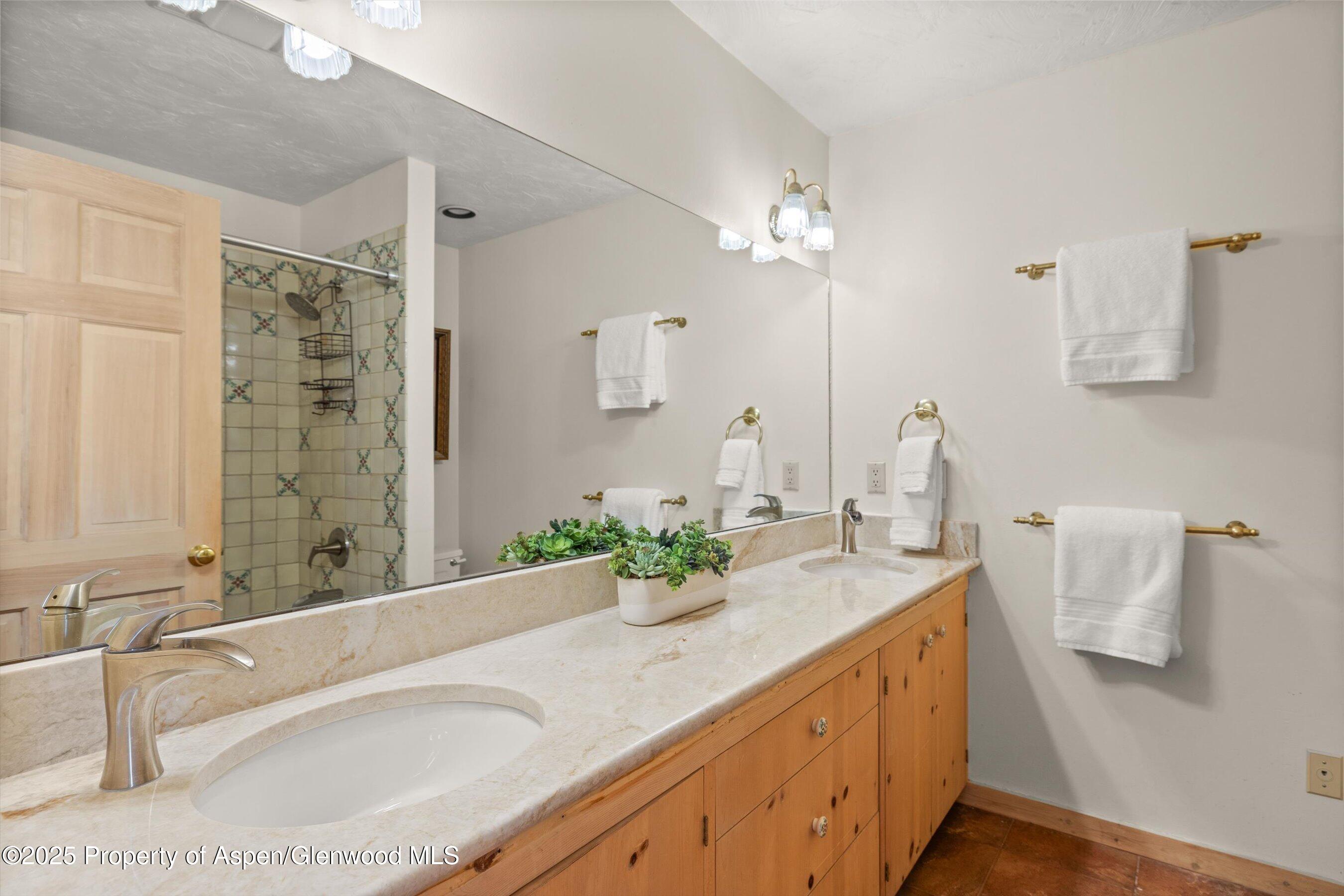 Bathroom vanity with a cream stone counter, wood cabinets, large mirror, and tiled shower.