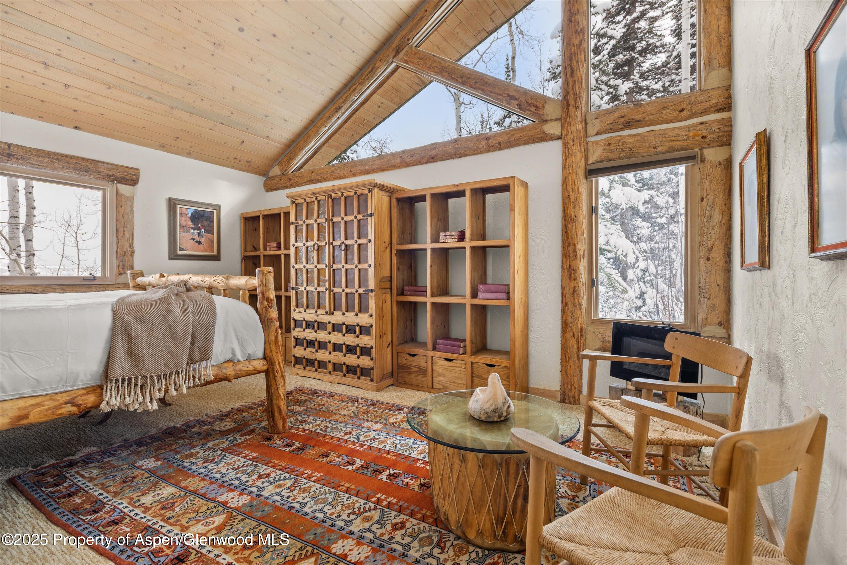 Rustic bedroom and sitting area with a log bed, timber beams, and windows overlooking snowy trees.