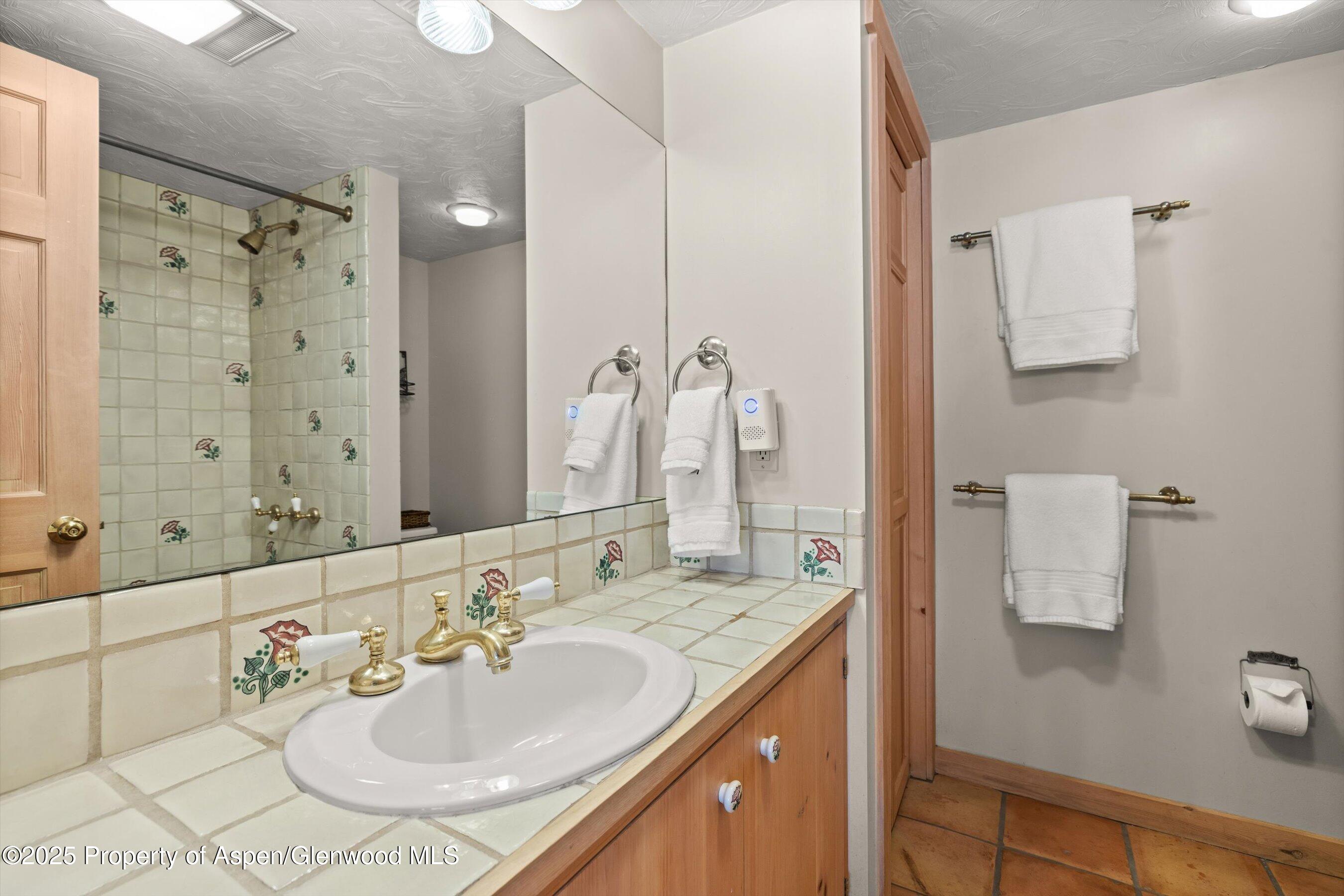 Bathroom with floral-accented tile vanity, oval sink, brass fixtures, and tiled shower.