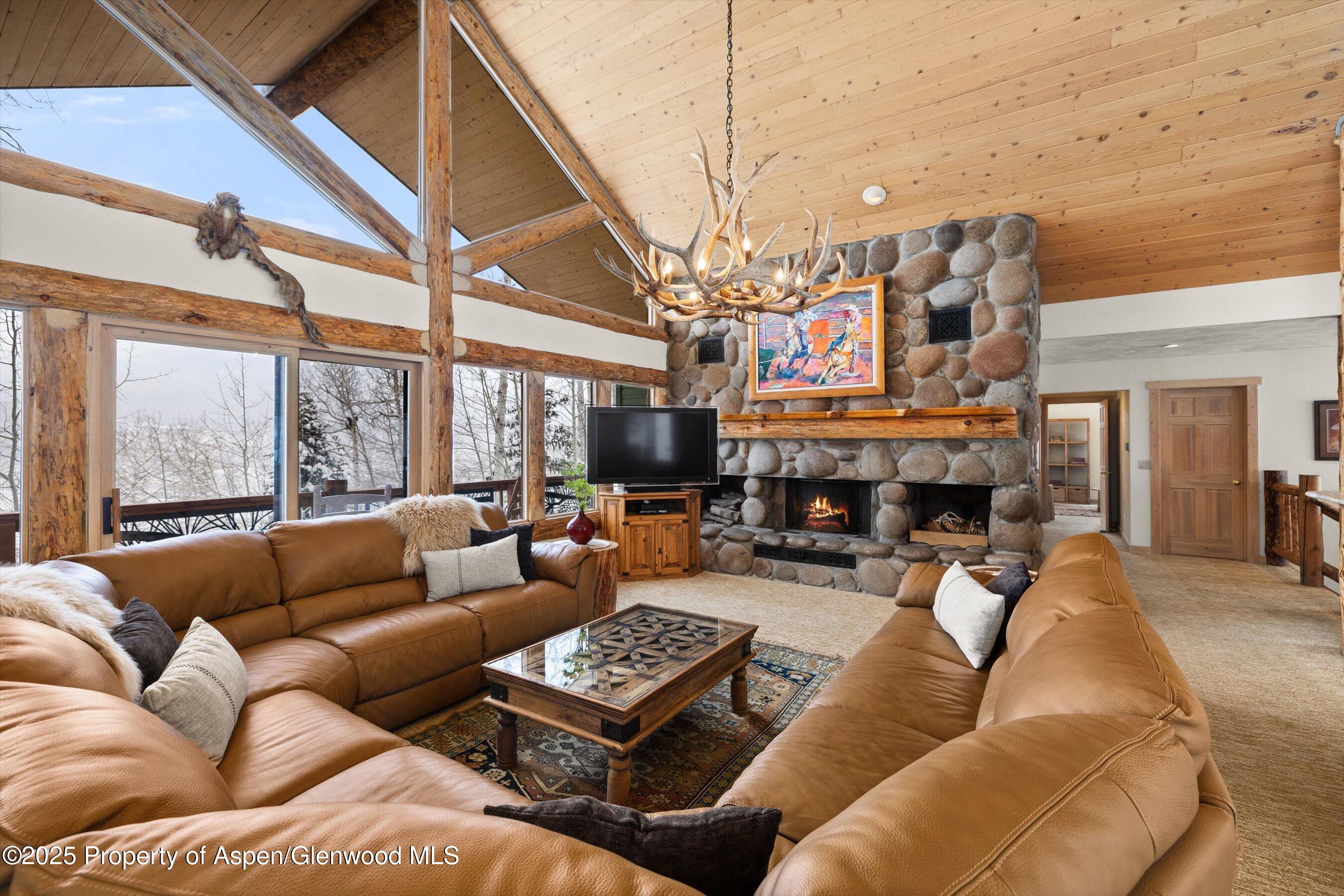 Mountain living room with vaulted timber ceiling, stone fireplace, antler chandelier, and windows overlooking snowy trees.