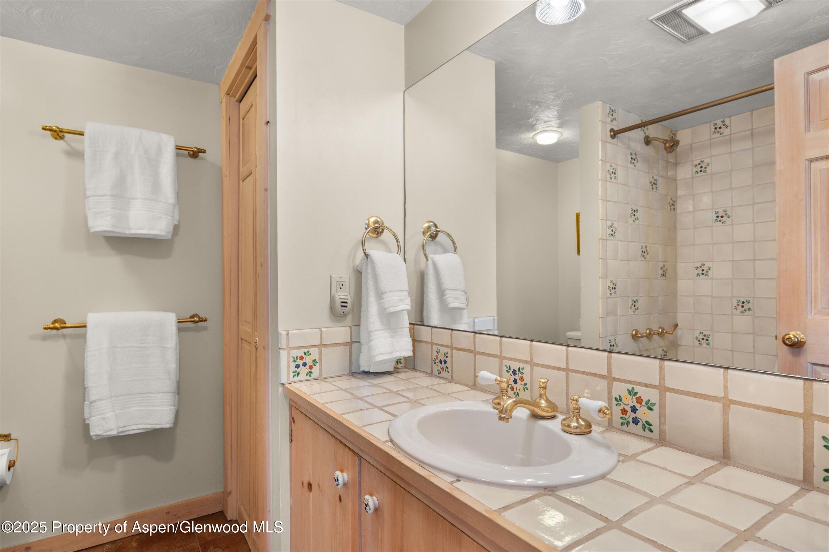 Bathroom vanity and tiled shower with floral accents, brass fixtures, and white towels.
