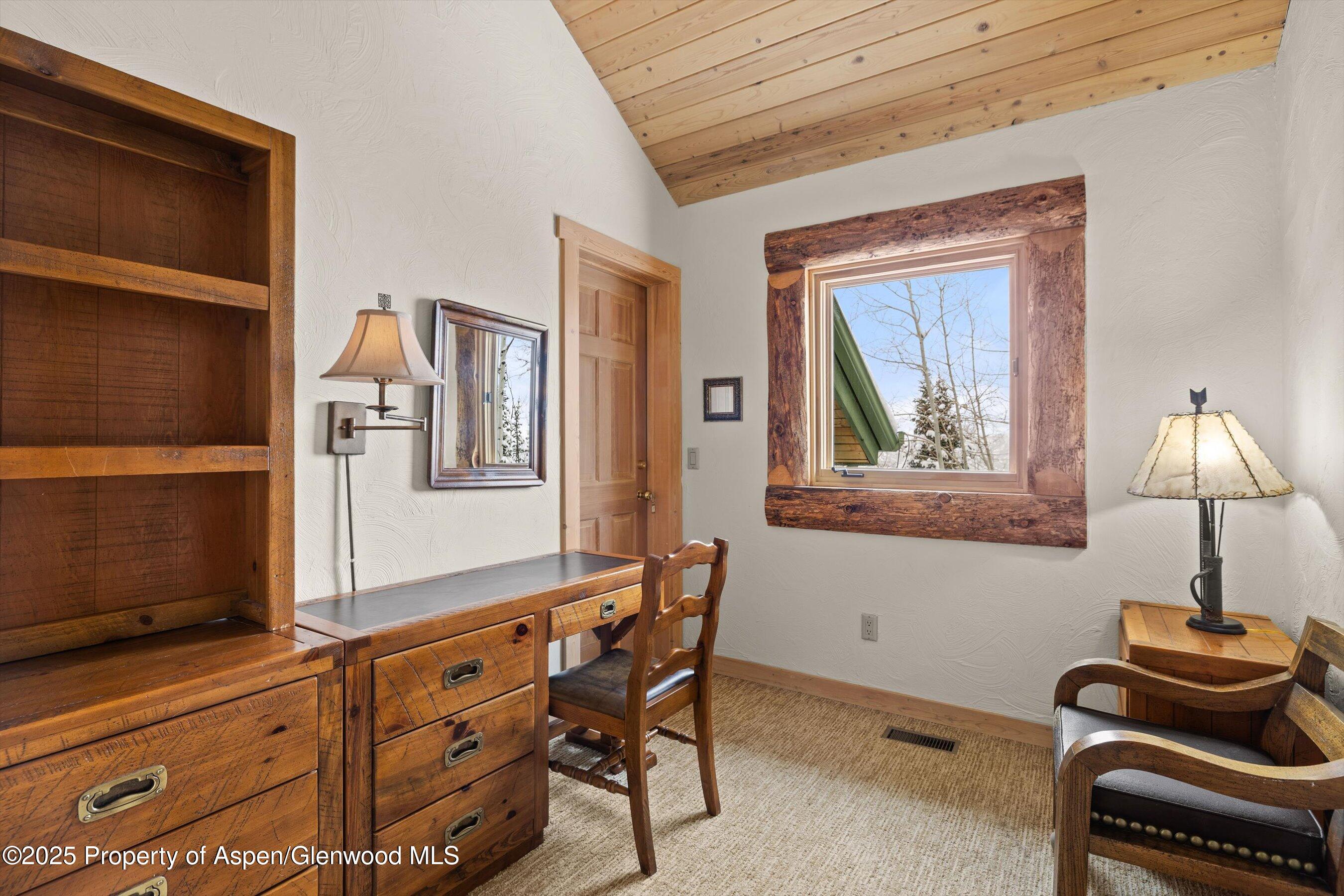 Furnished home study with rustic wooden desk, built-in shelves, vaulted ceiling, and snow visible through a timber-framed window.