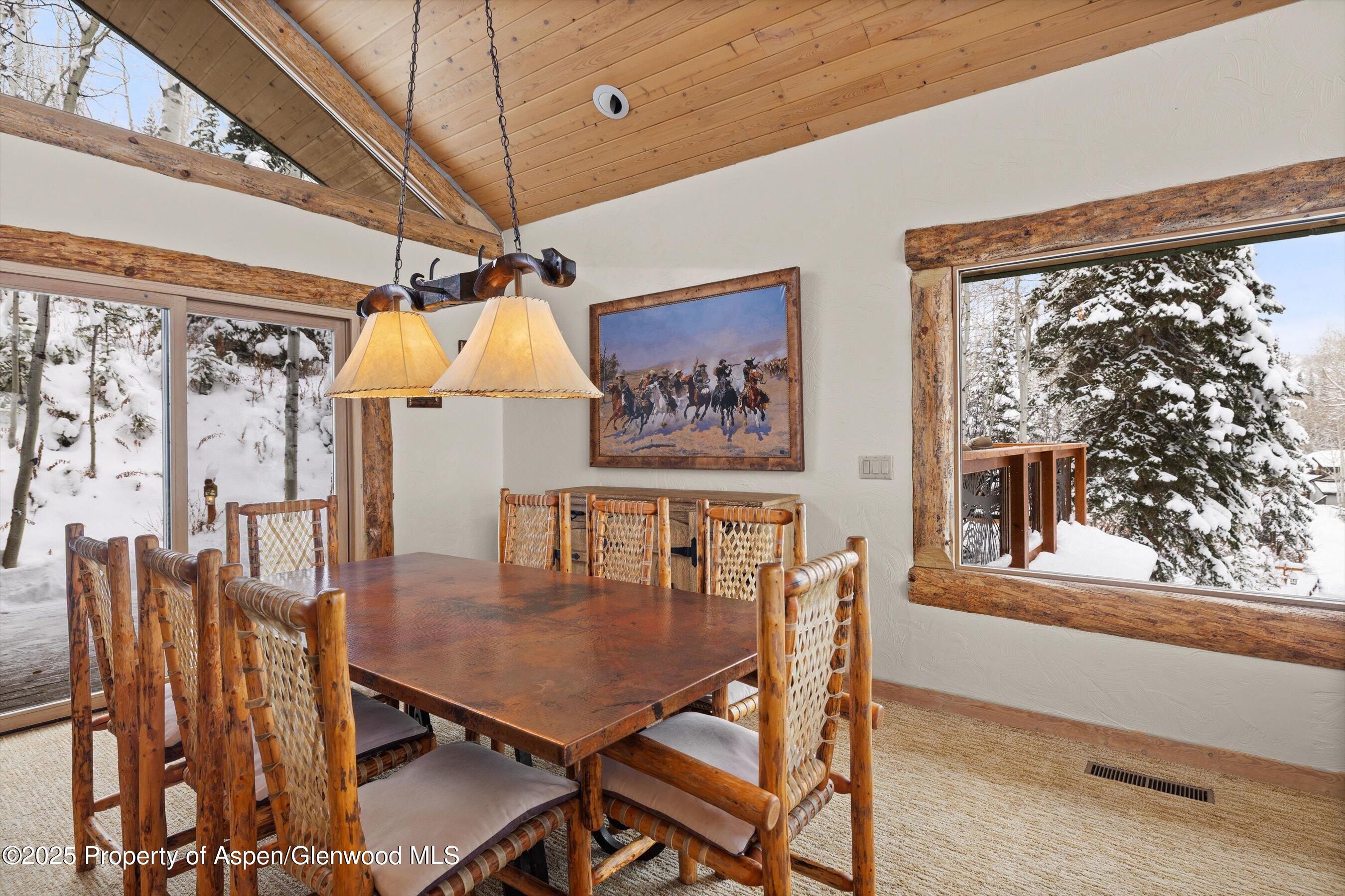 Dining room with rustic timber ceiling, woven wooden chairs, and snow-covered trees outside large windows.