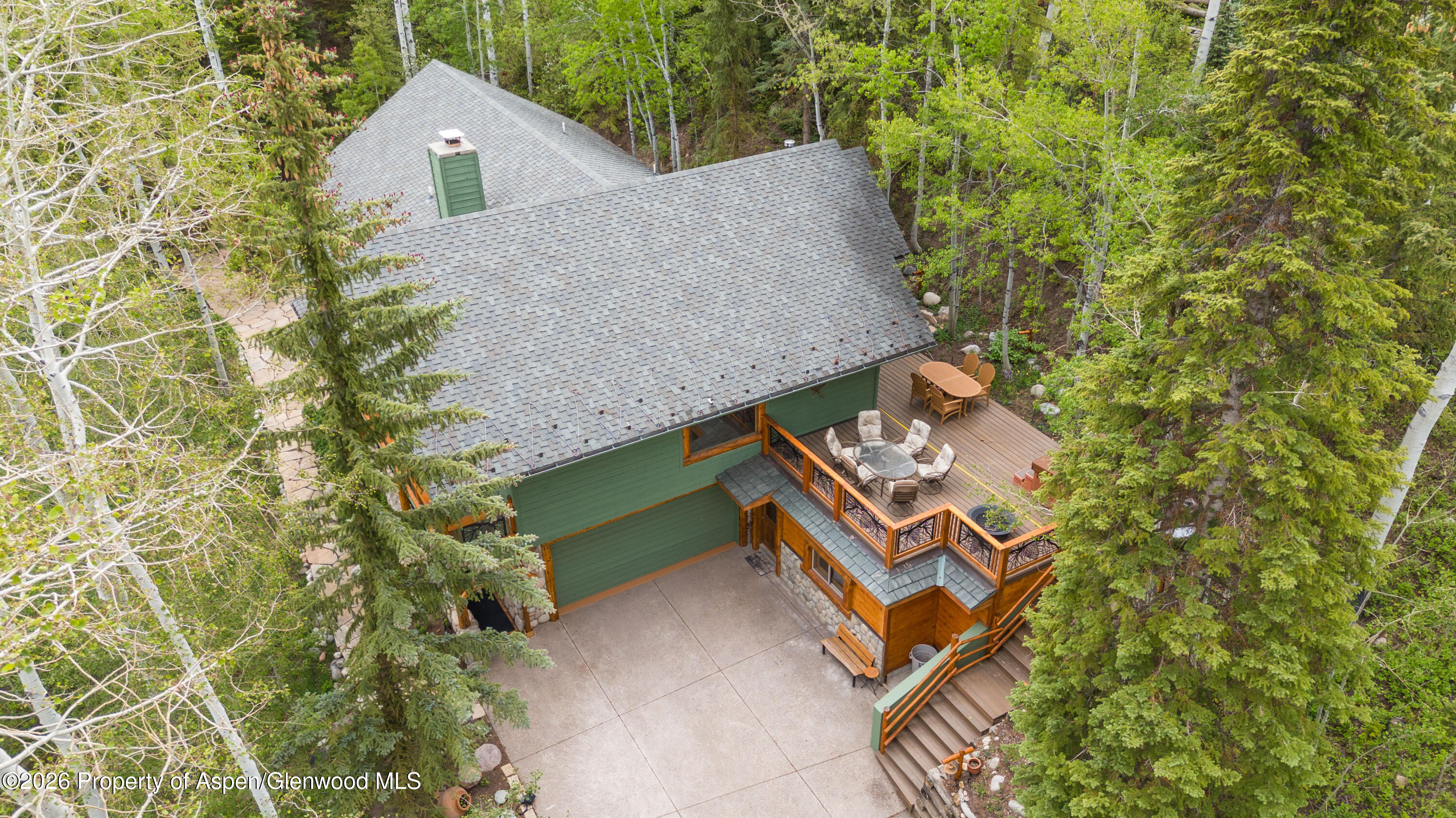 Aerial view of a wooded home with a furnished wraparound deck and paved driveway.