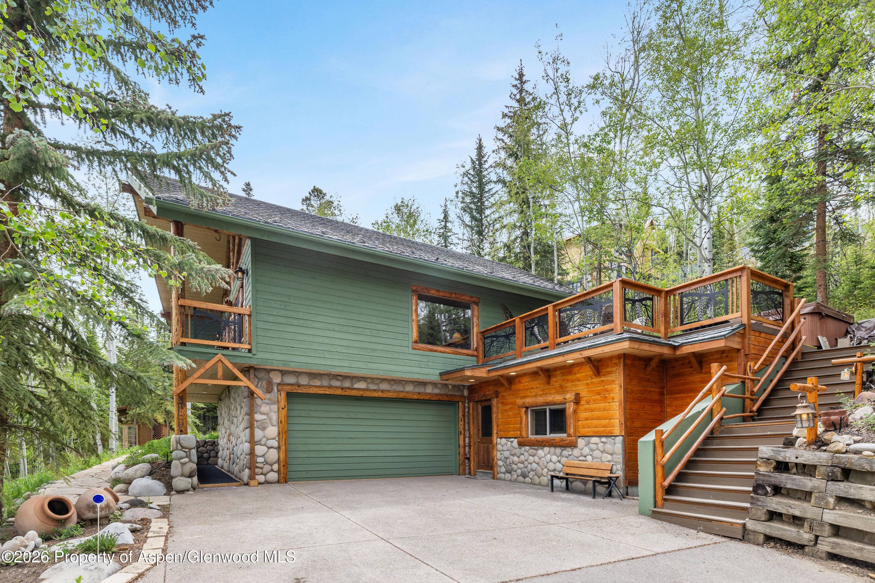 Mountain home with green siding, stone accents, and a raised wooden deck among mature trees.