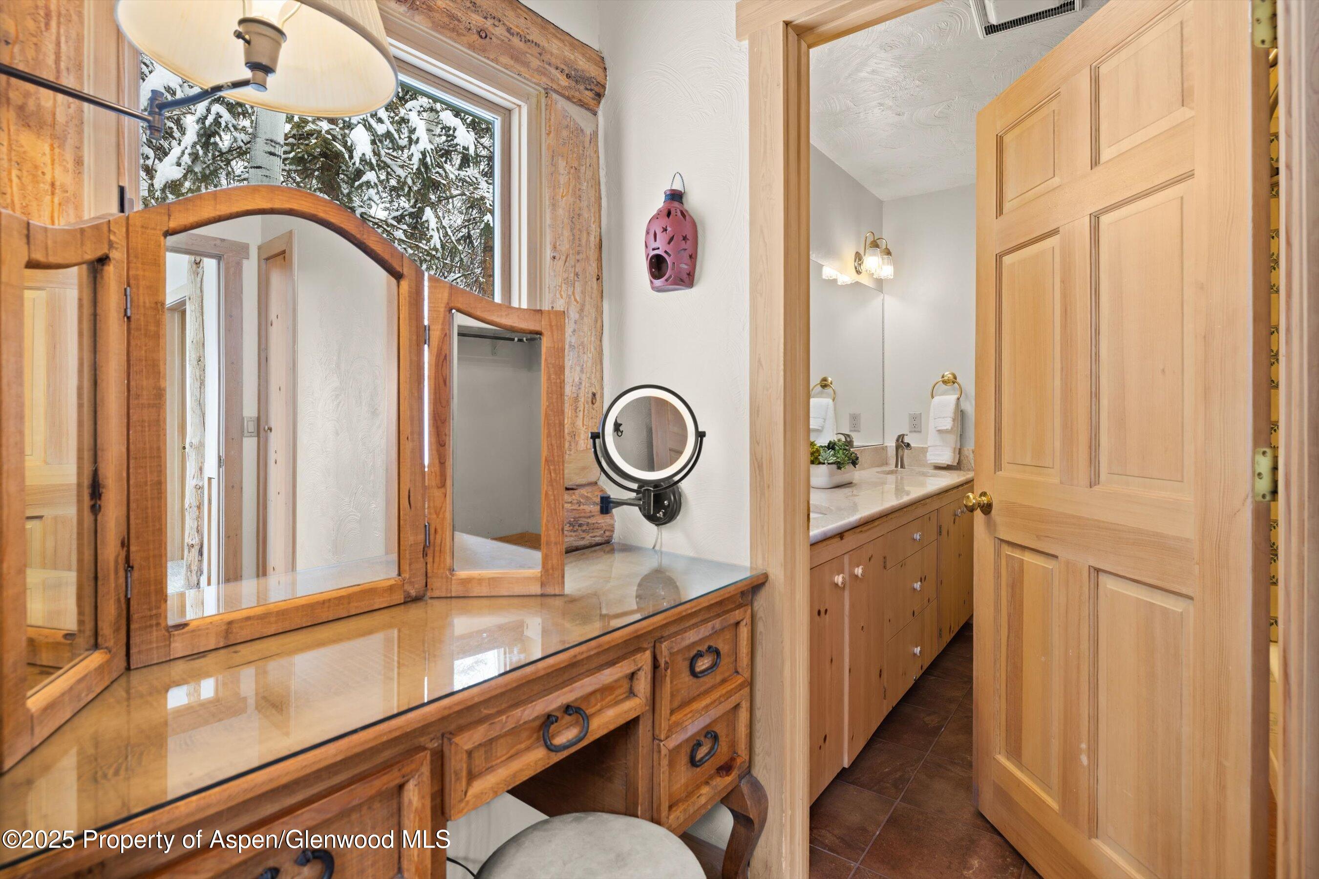 Rustic wood vanity beside a bathroom with a long countertop and sink; snowy evergreens show through the window.