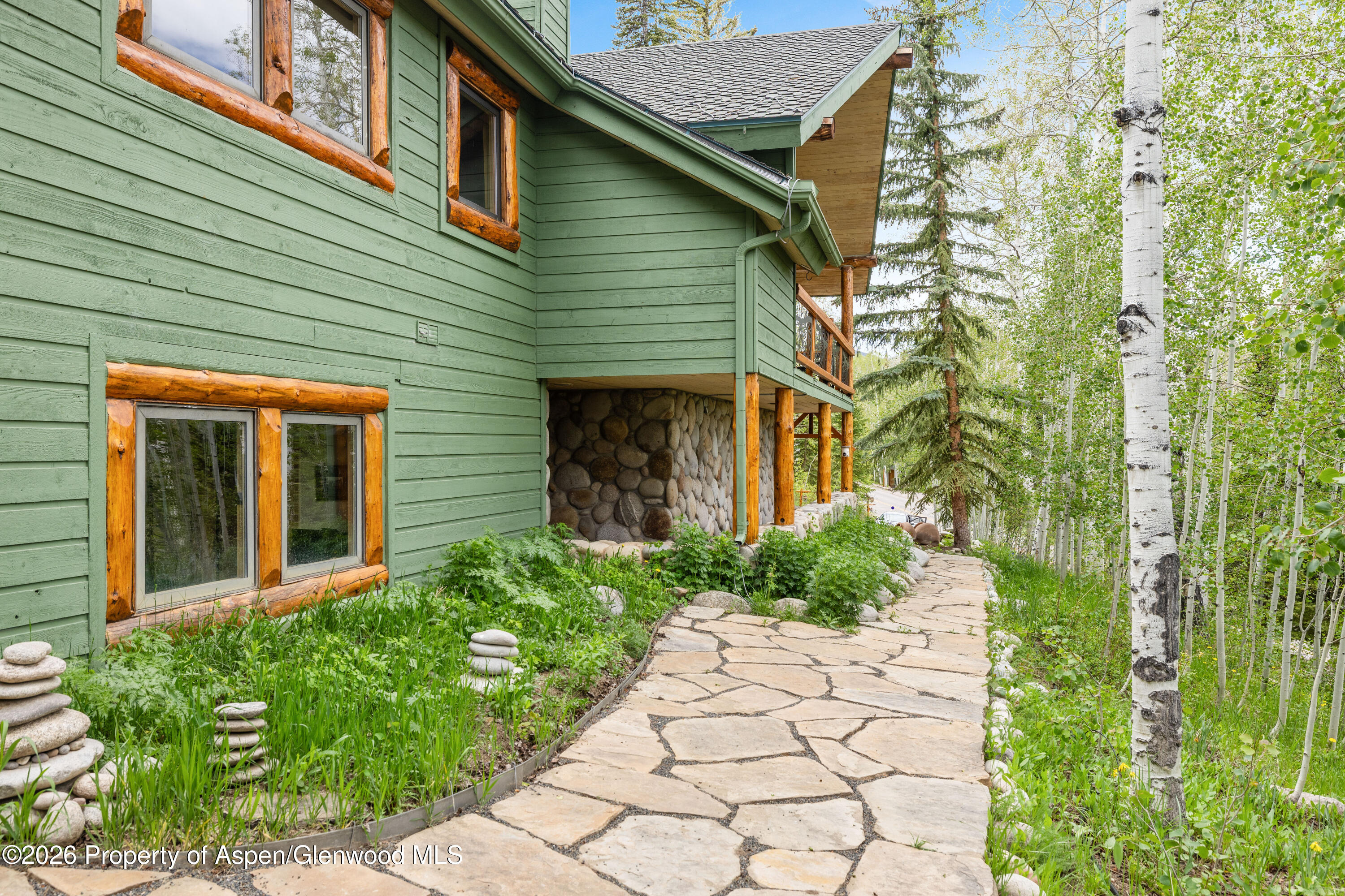 Green mountain home with timber-trimmed windows beside a flagstone path through aspen trees.