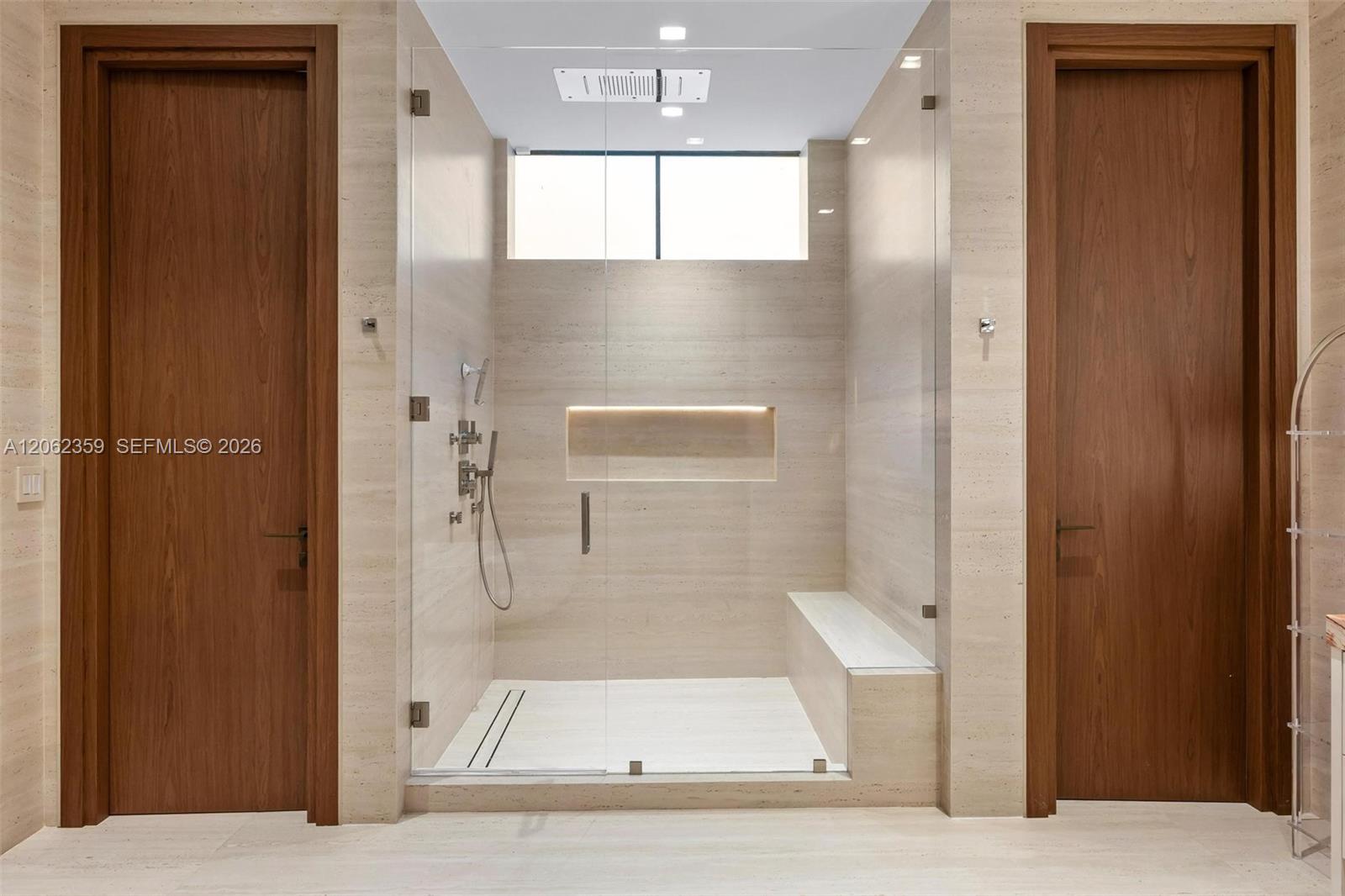 Walk-in shower with pale stone tile, glass enclosure, illuminated niche, and built-in bench.