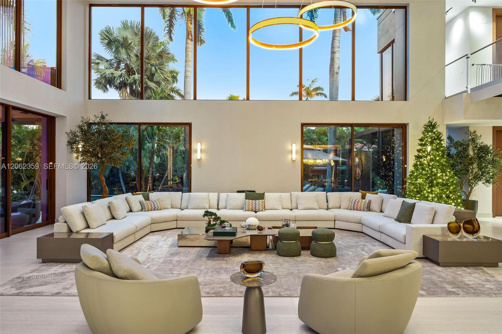 Double-height living room with a curved cream sectional, ring chandelier, and windows overlooking palms.