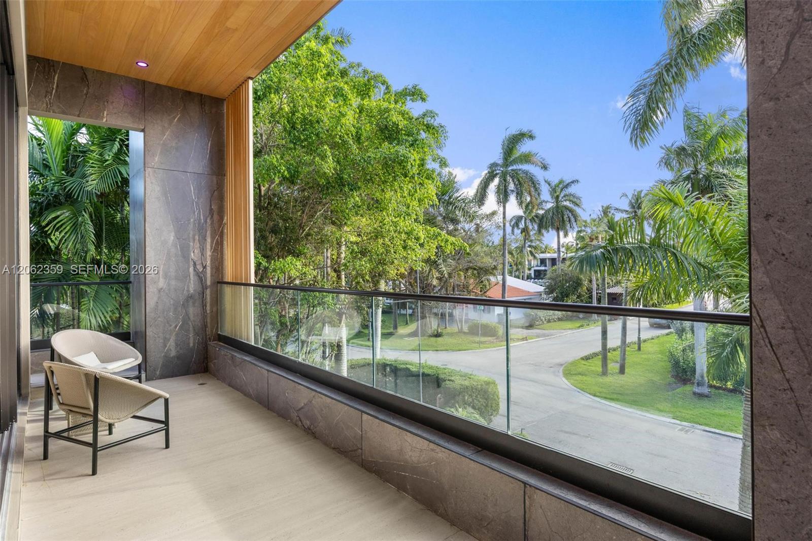 Covered balcony with woven chairs and glass railing overlooking a palm-lined street.