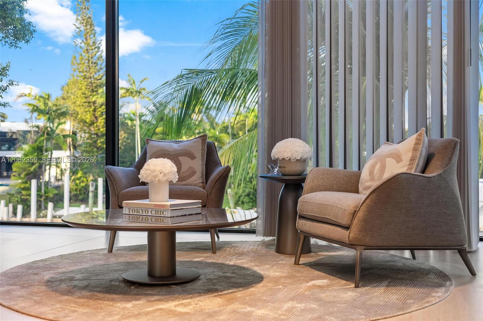 Living room with two upholstered armchairs and a round coffee table beside floor-to-ceiling windows overlooking palms.