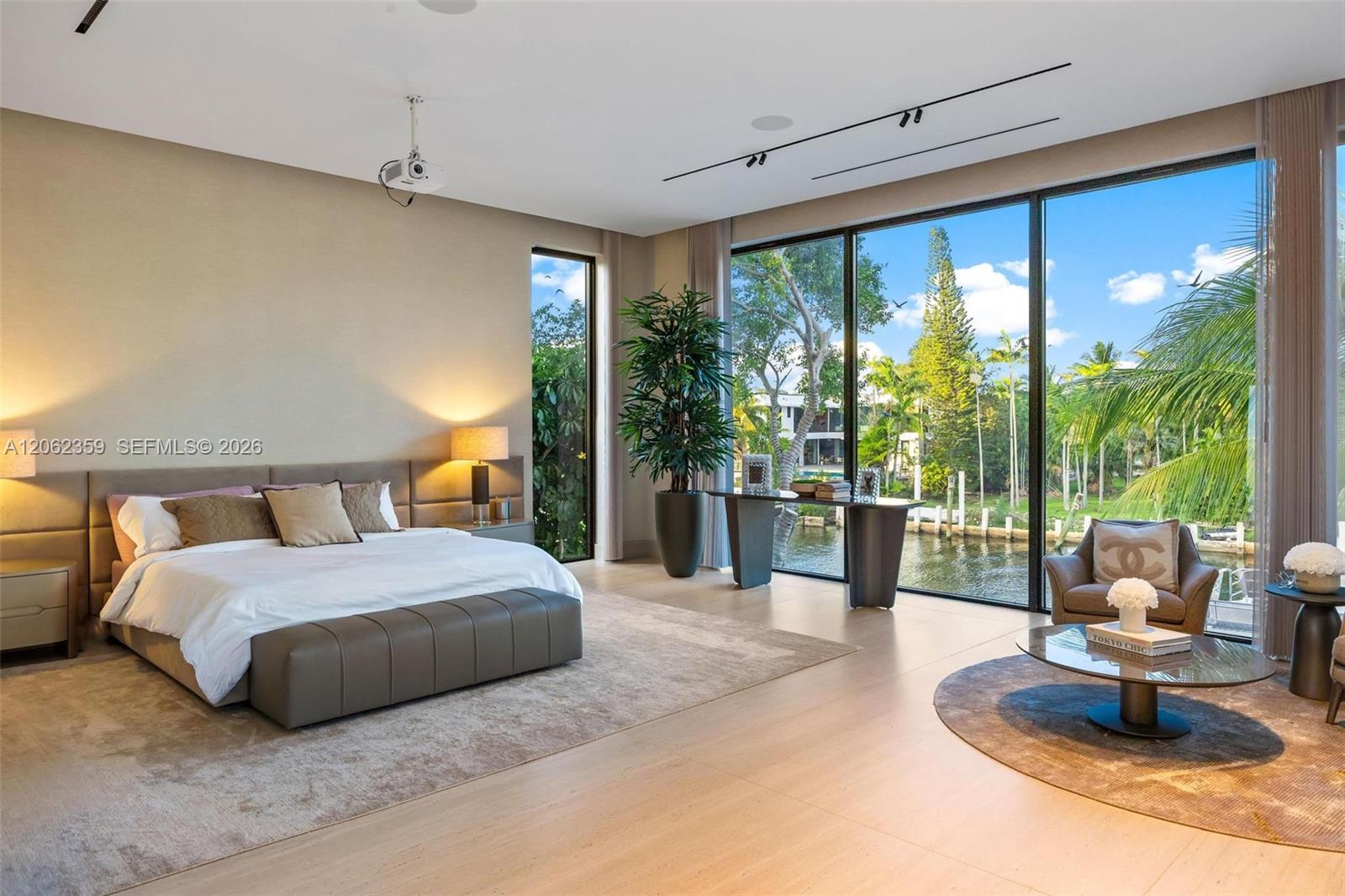 Modern bedroom with a large upholstered bed and floor-to-ceiling windows overlooking a tropical canal.