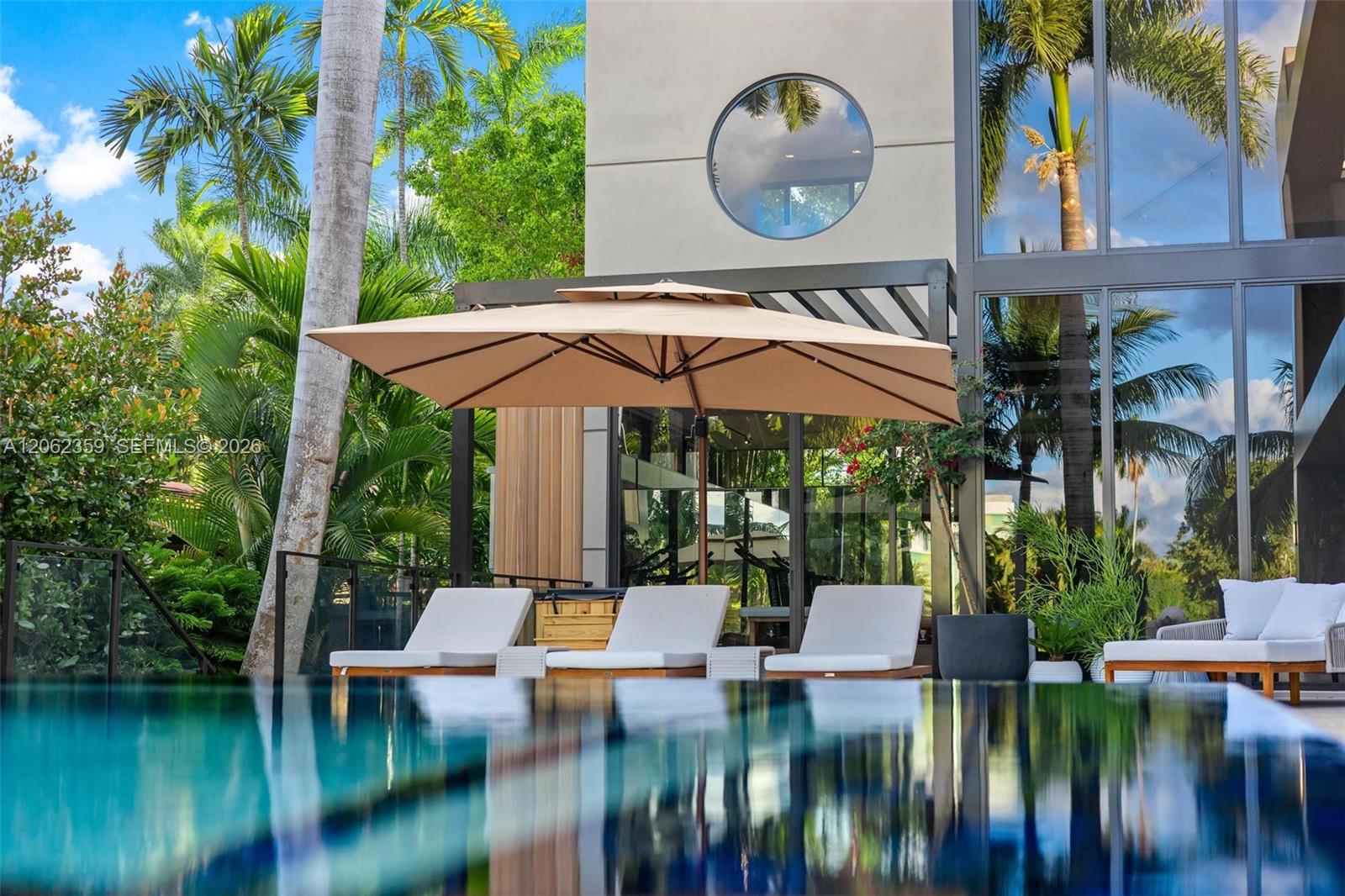 Modern home beside a reflective pool, with glass walls, tropical palms, and shaded patio seating.
