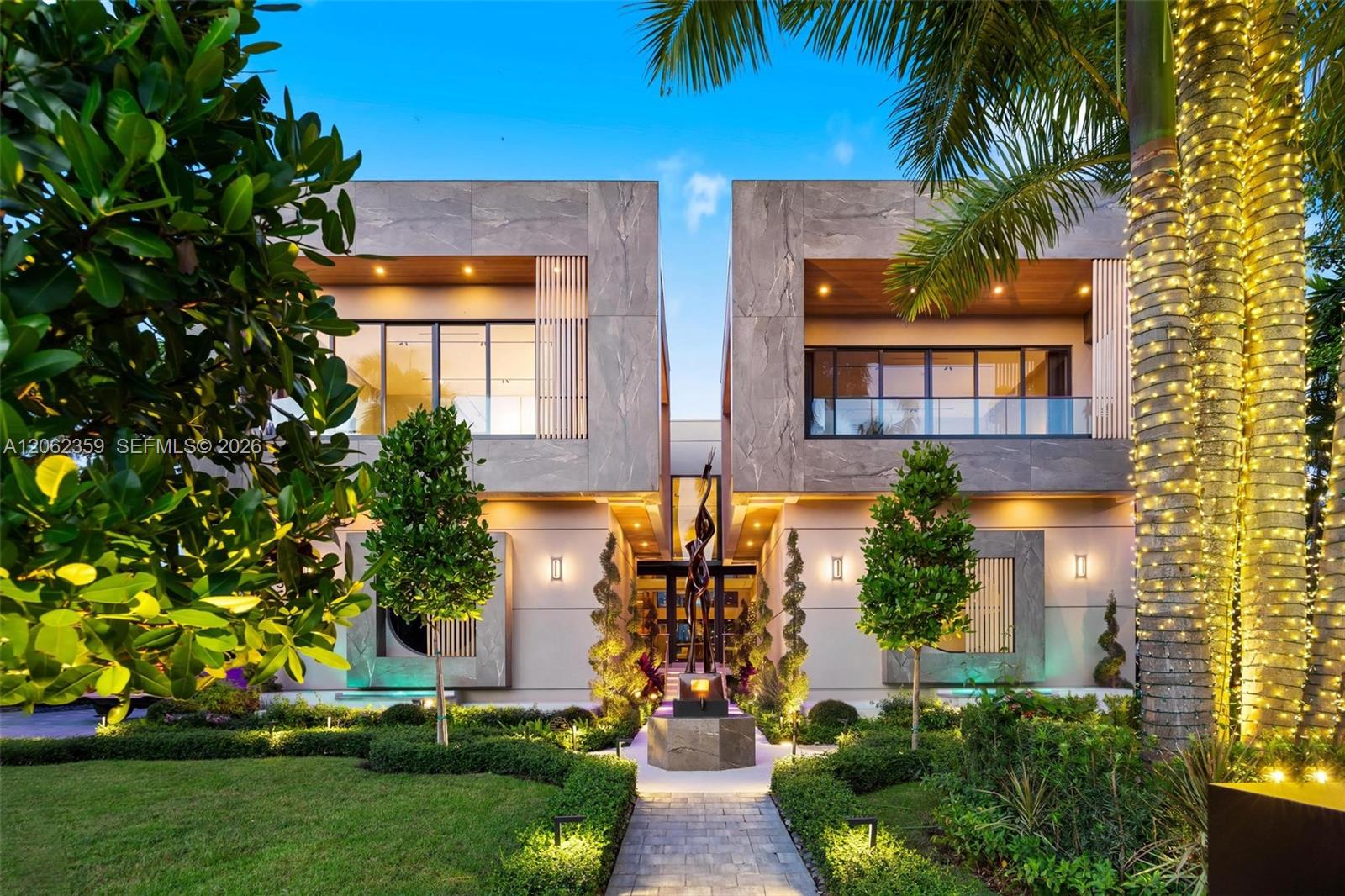 Modern two-story home with a sculptural entry walkway, lush landscaping, and illuminated palm trees.