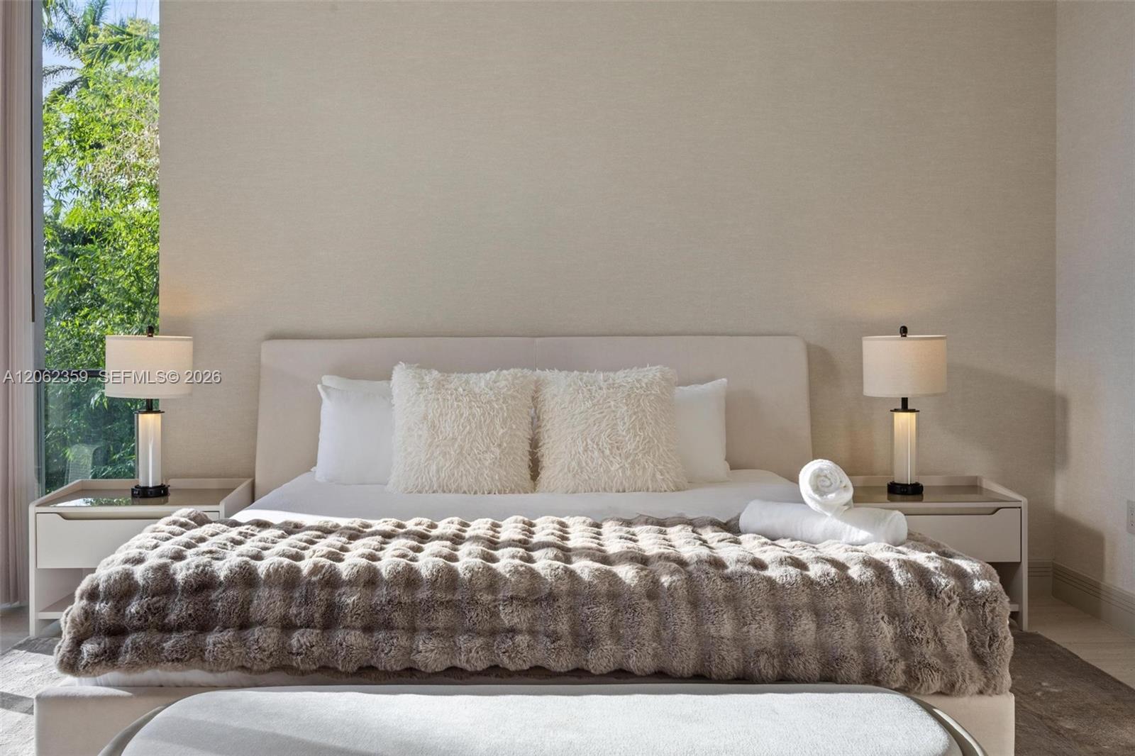 Bedroom with an upholstered bed, textured gray blanket, matching lamps, and a tall window overlooking greenery.