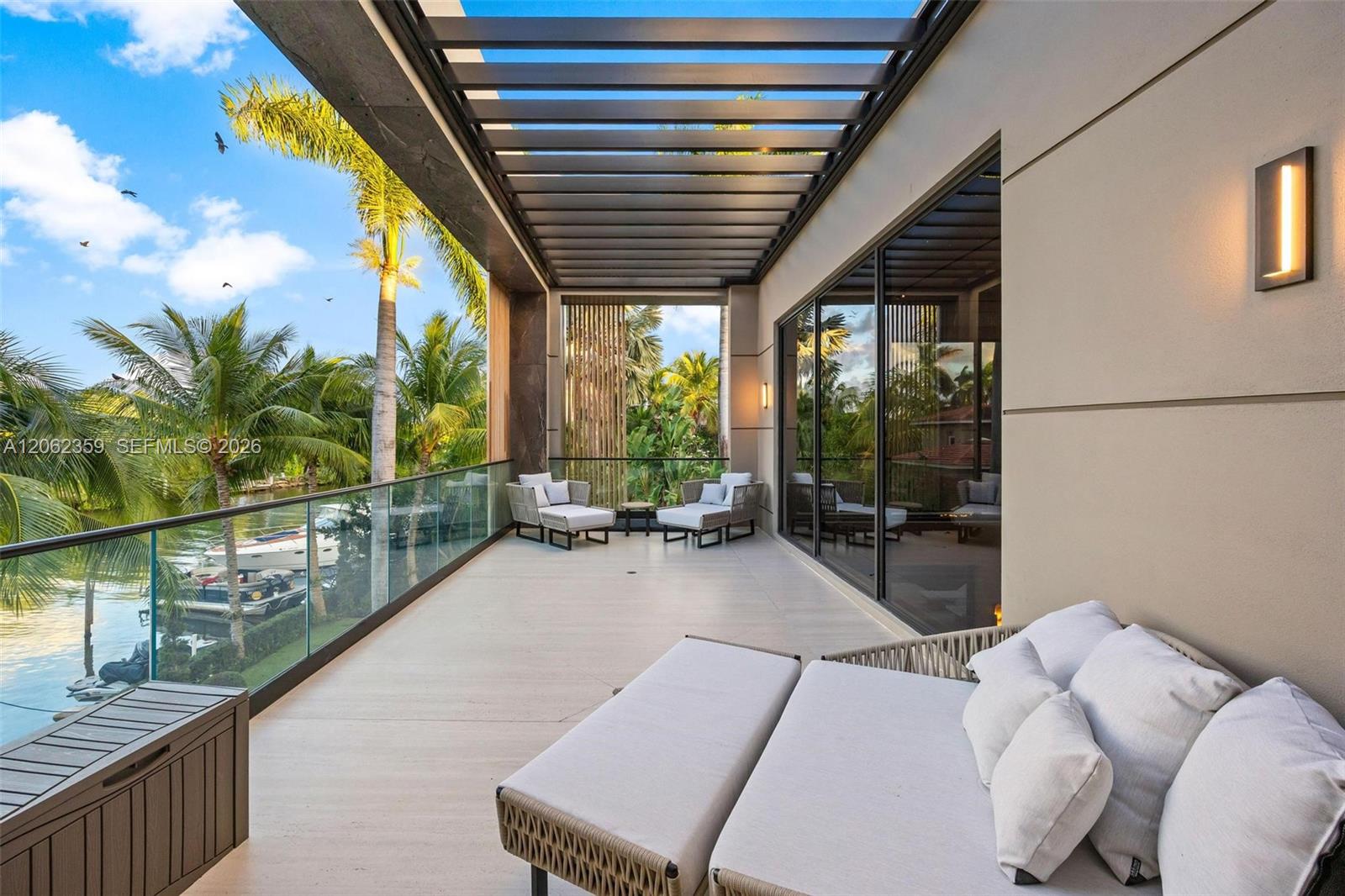 Covered balcony with glass railing, canal view, palm trees, and contemporary outdoor lounge seating.