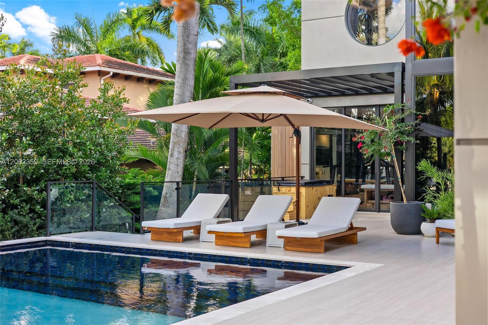 Modern home pool patio with three loungers under an umbrella and tropical landscaping.