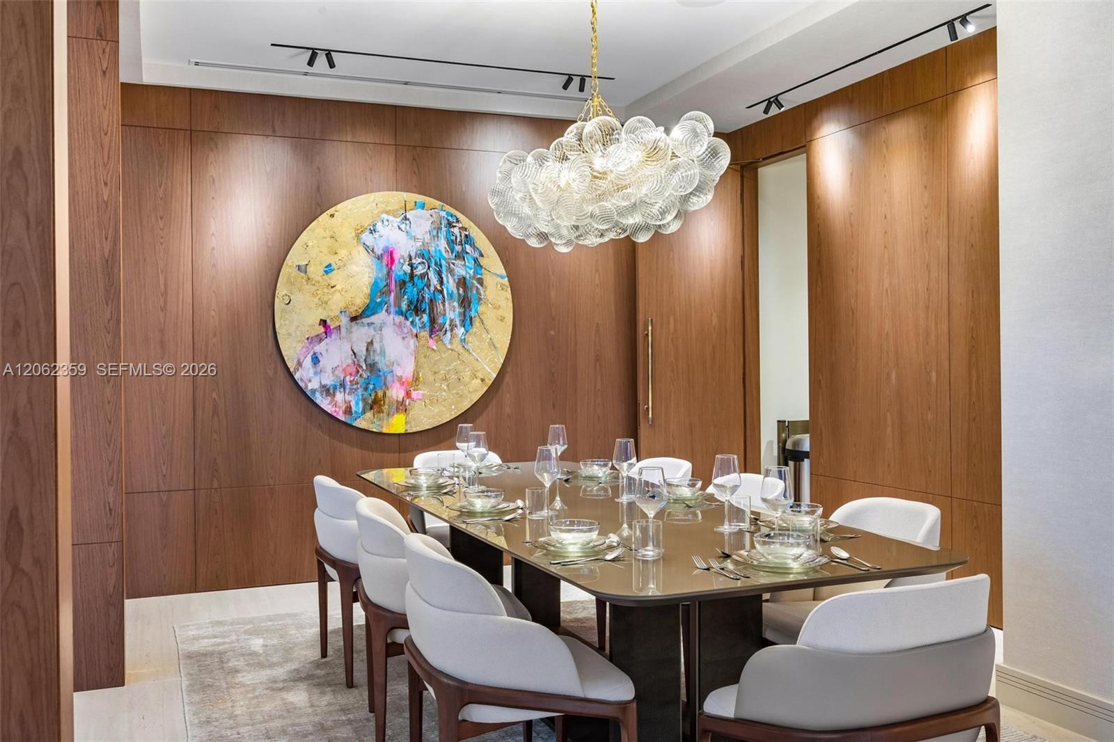 Dining room with a set glass table, white chairs, walnut paneling, and a sculptural glass chandelier.