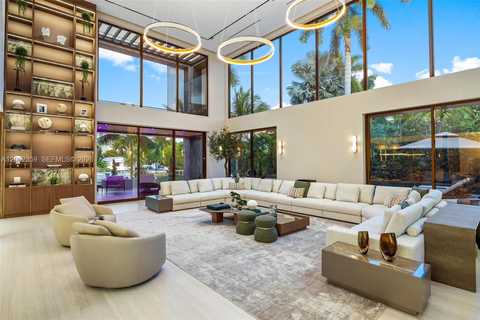 Double-height living room with a cream sectional, tall display shelving, and floor-to-ceiling windows overlooking palms.