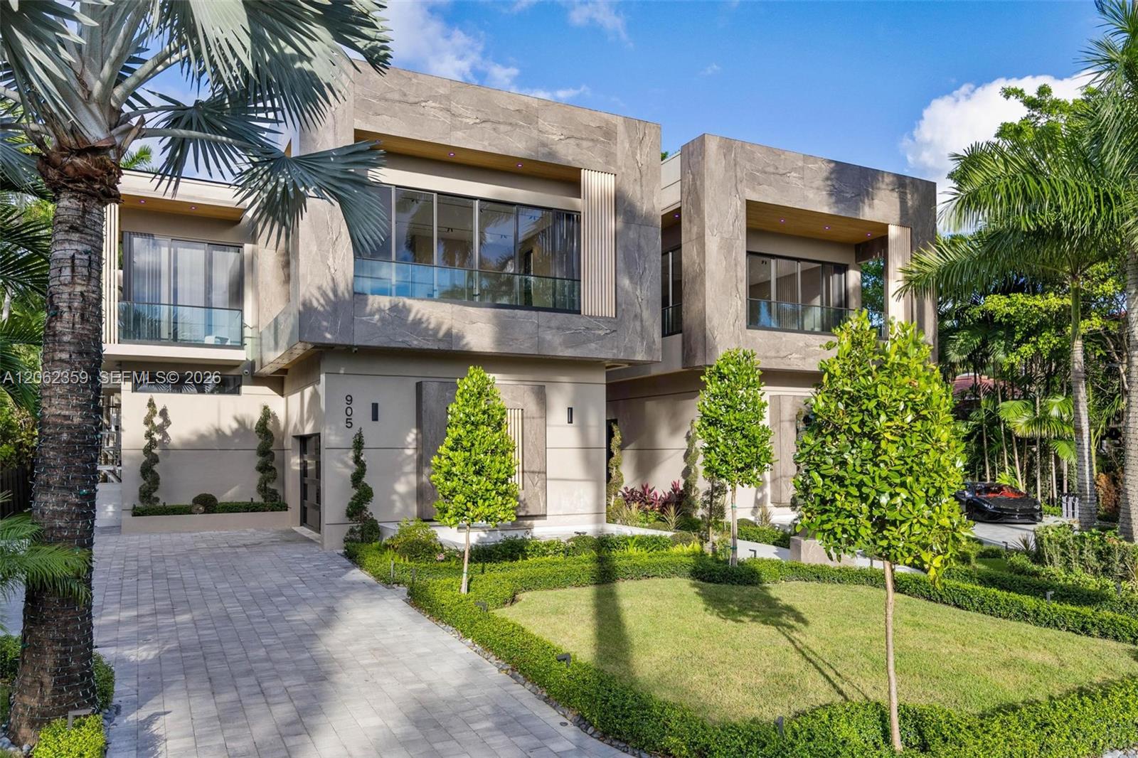 Modern two-story home with a stone-look façade, tropical landscaping, and a broad paved walkway.