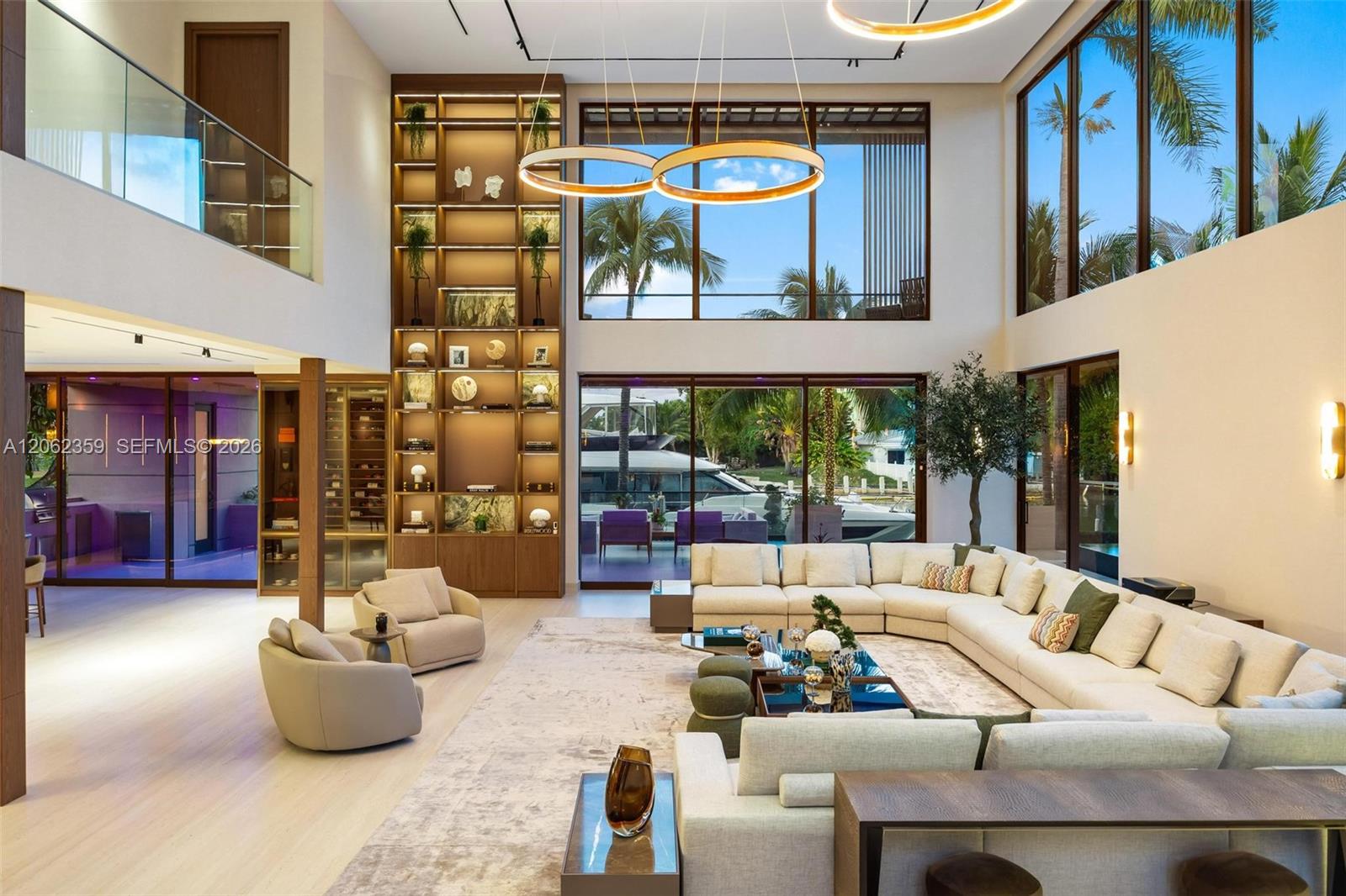 Double-height living room with cream sofas, illuminated shelving, and windows overlooking palms and a yacht.