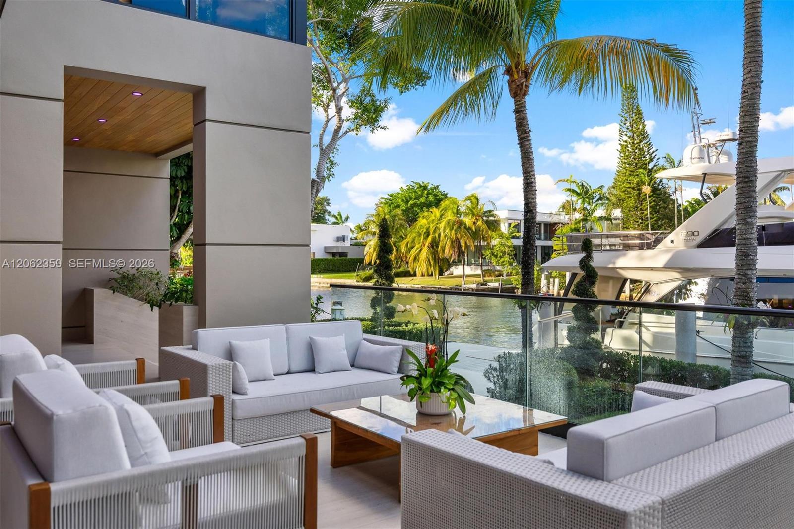 Waterfront patio with white seating, a glass railing, palm trees, and a yacht on the canal.