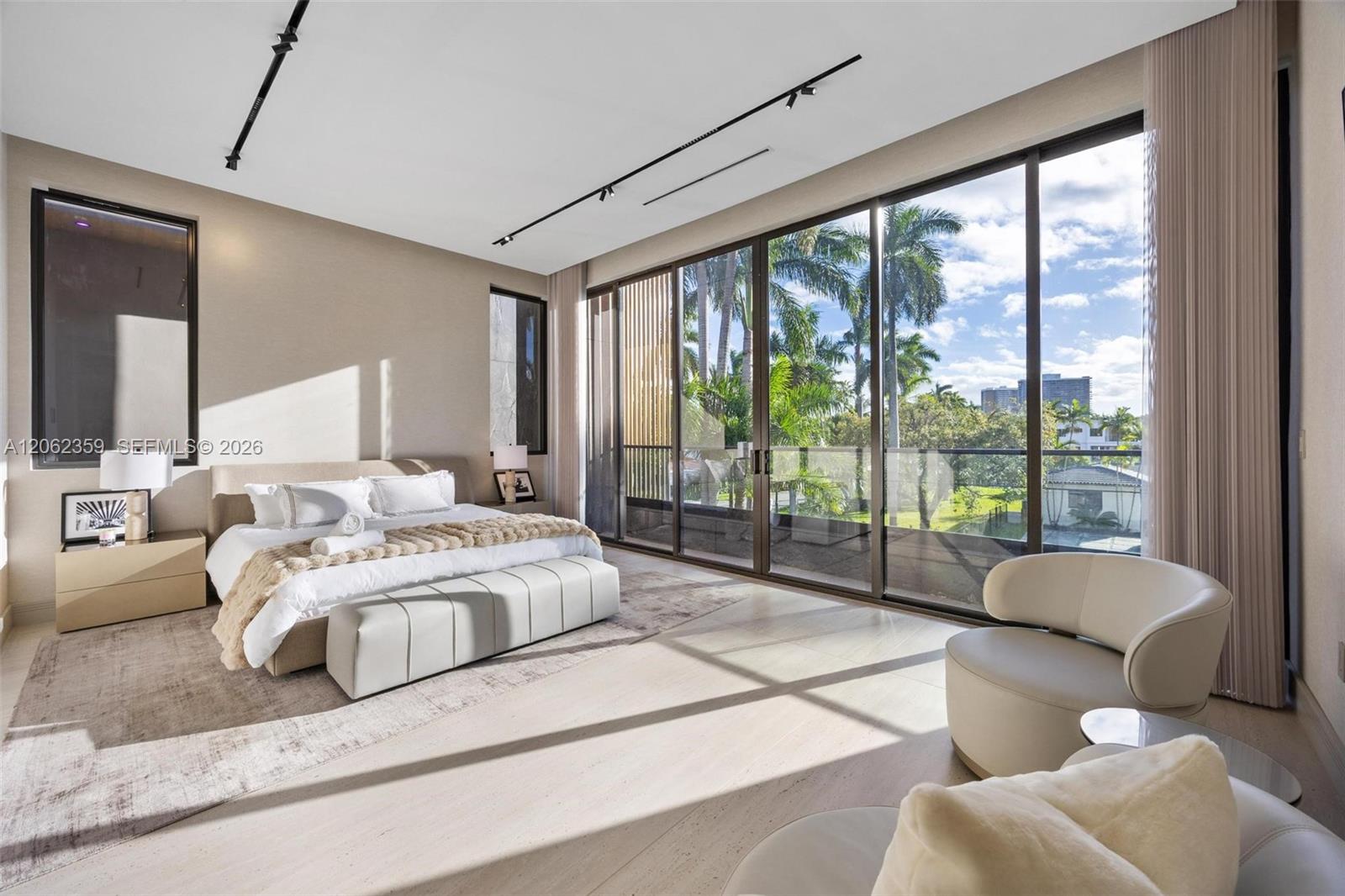 Modern bedroom with a low upholstered bed and floor-to-ceiling windows overlooking palm trees.