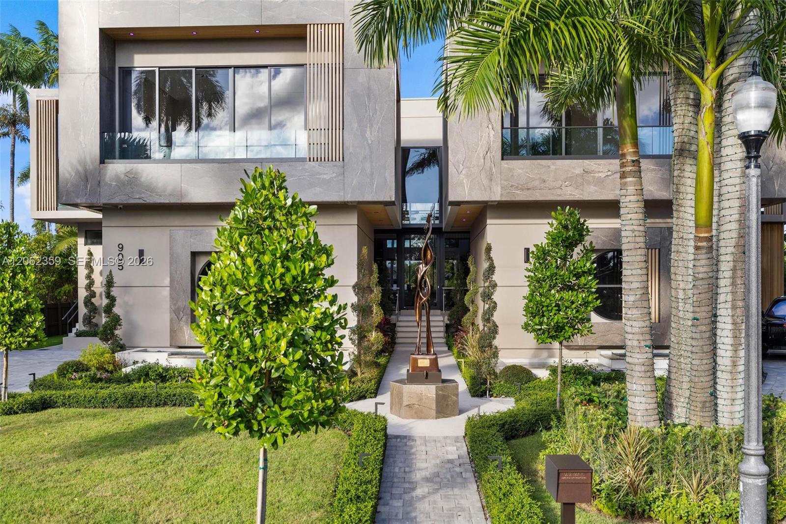 Modern stone-clad home with expansive windows, tropical landscaping, and a bronze sculpture at the entrance.