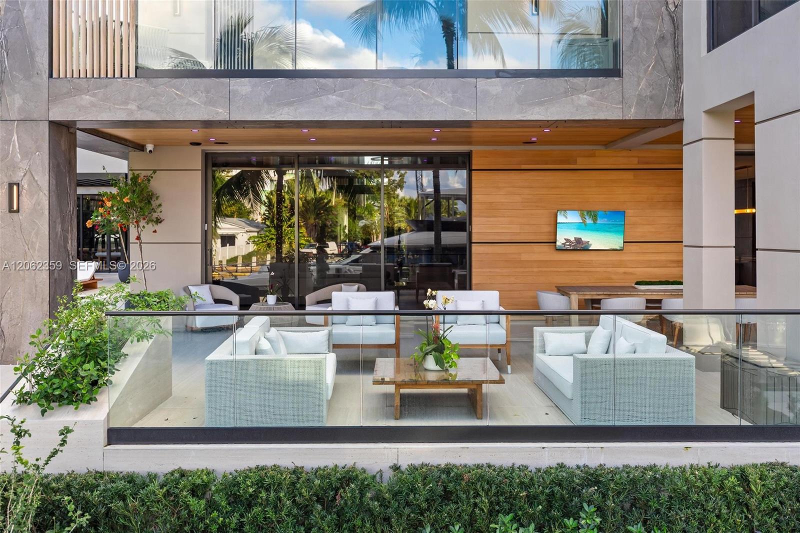 Covered patio with pale lounge seating, glass railing, wood-paneled wall, and tropical greenery.