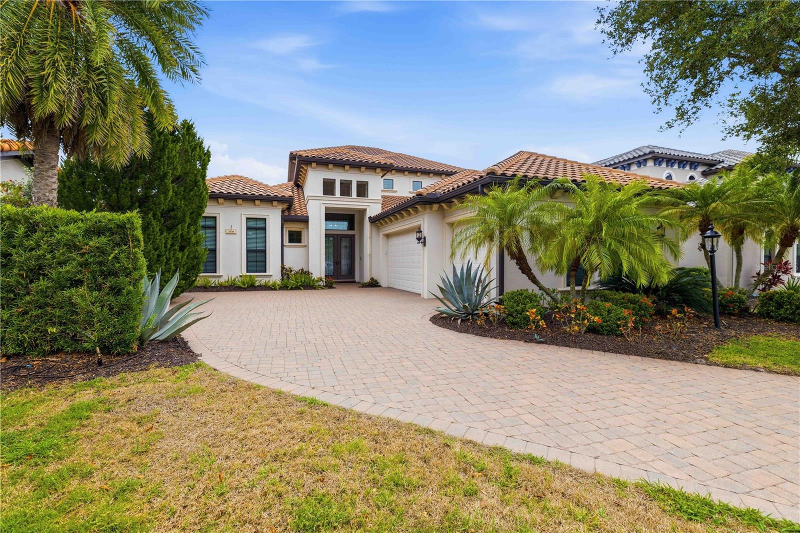 Single-story stucco home with a tile roof, curved paver driveway, and tropical landscaping.