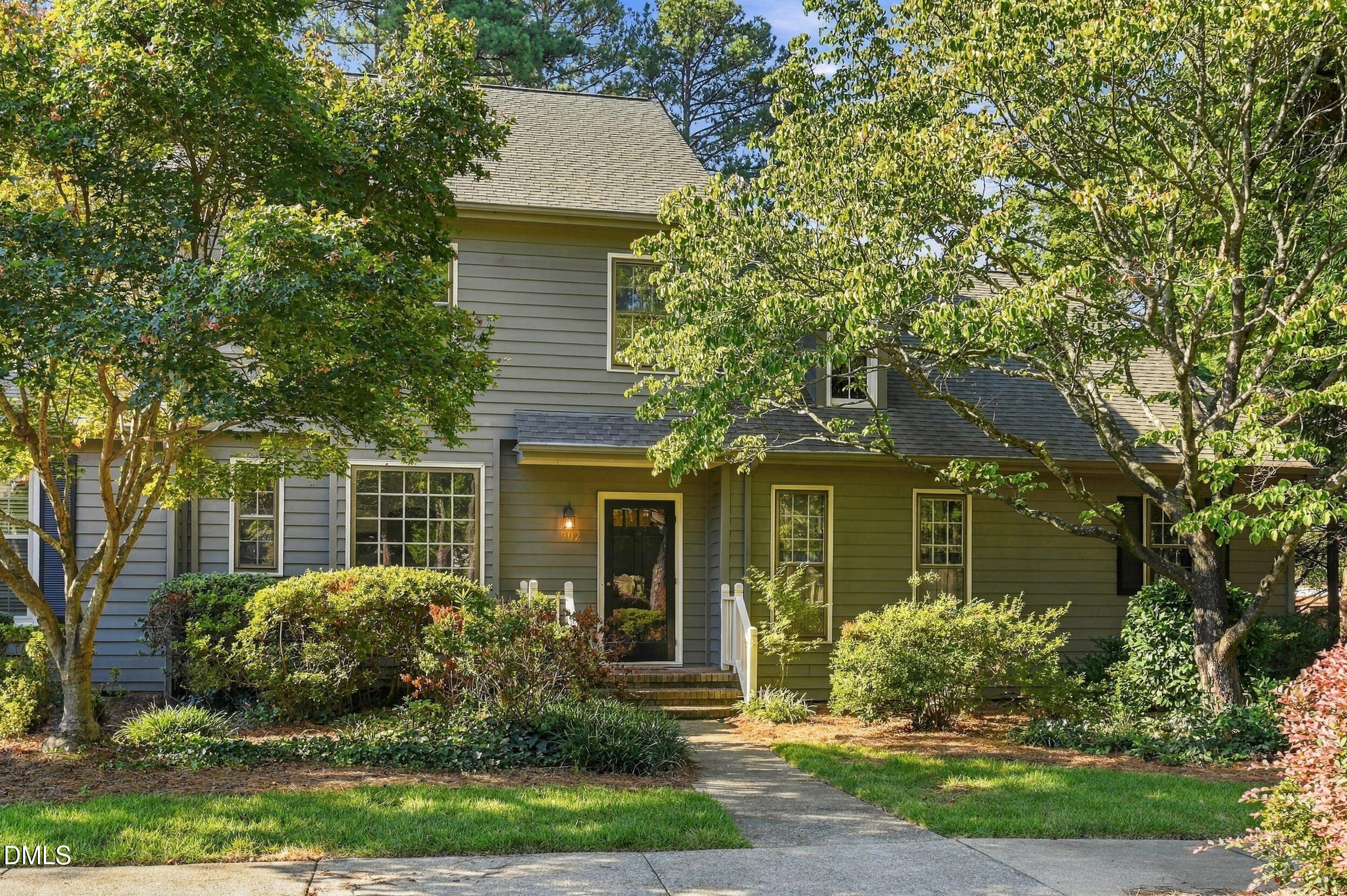 Two-story gray-sided home framed by mature trees, with a landscaped front yard and white entry railings.