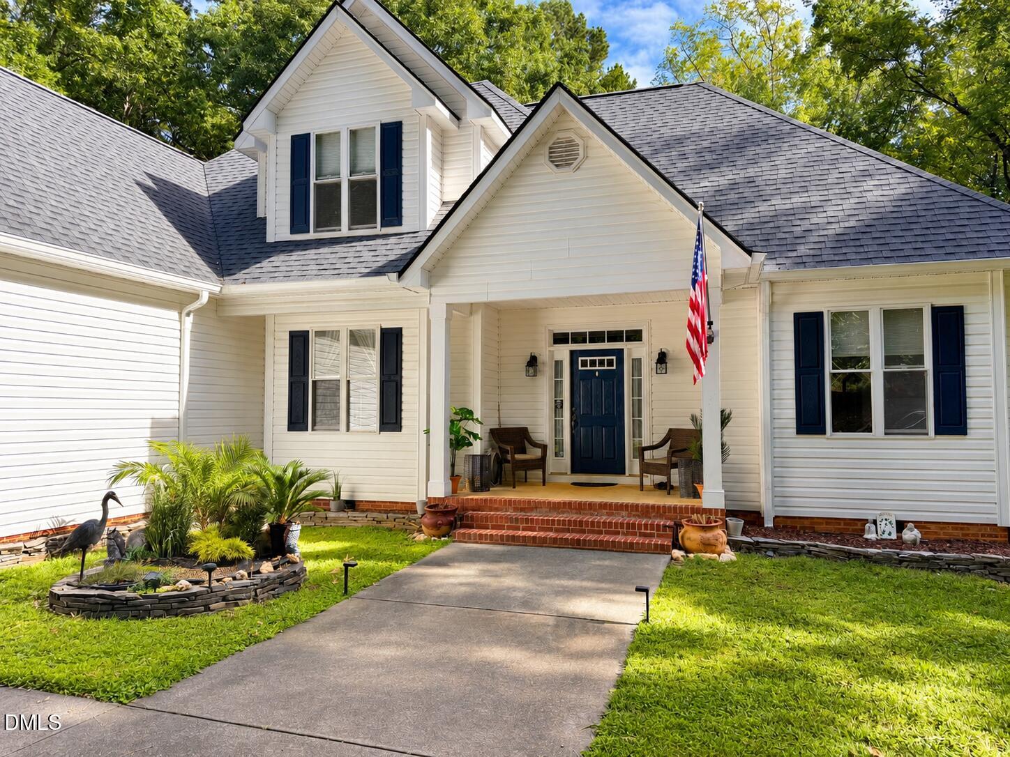 White two-story home with blue shutters, a covered porch, brick steps, and landscaped gardens.