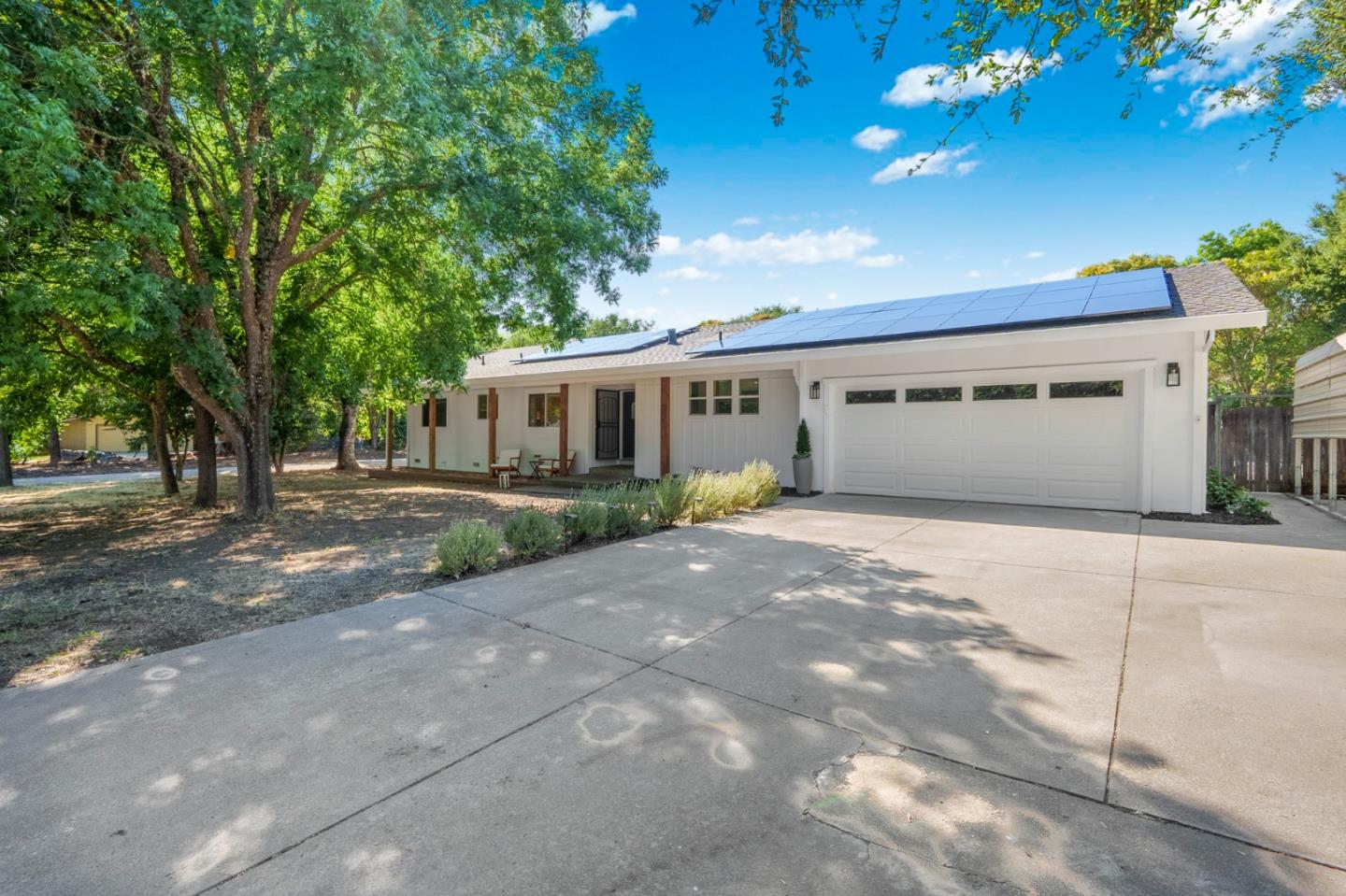 Single-story white house with rooftop solar panels, a two-car garage, and mature shade trees.