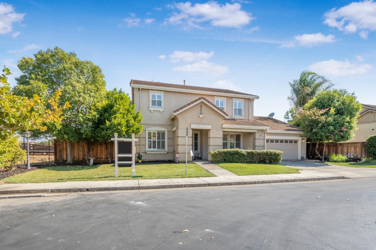 Two-story beige stucco house with an attached garage, front lawn, and mature trees.
