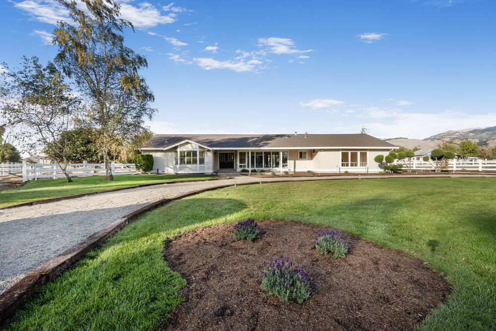 Single-story ranch home beyond a curved gravel drive and landscaped lawn, with white fencing and distant hills.