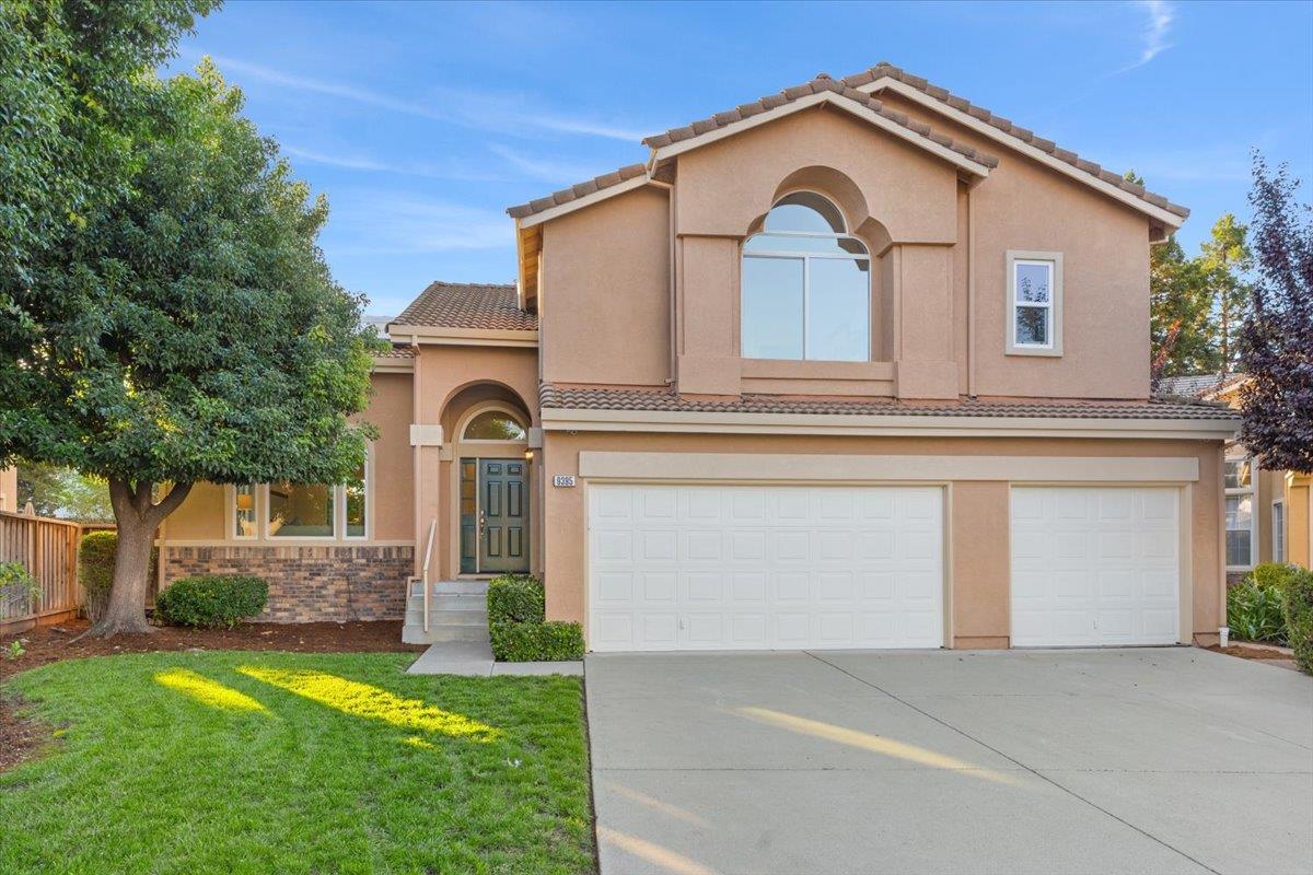 Two-story tan stucco house with arched windows, a double garage, and a green front lawn.