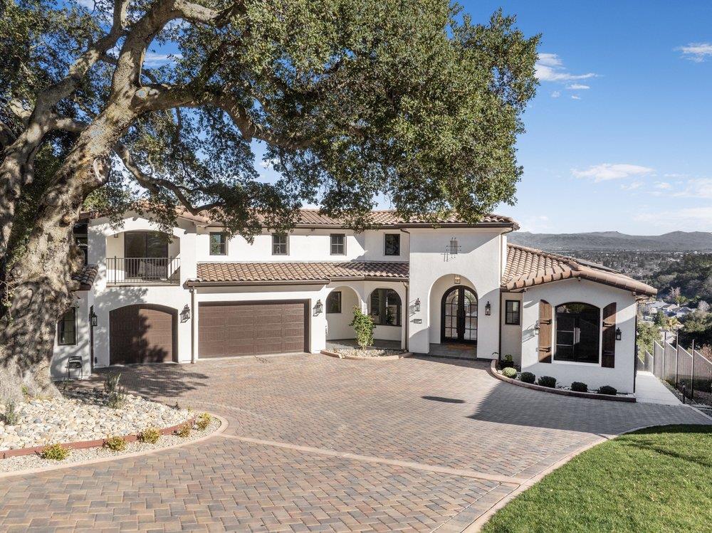 Two-story stucco home with a tiled roof, arched entry, brick-paver driveway, and mature oak tree.